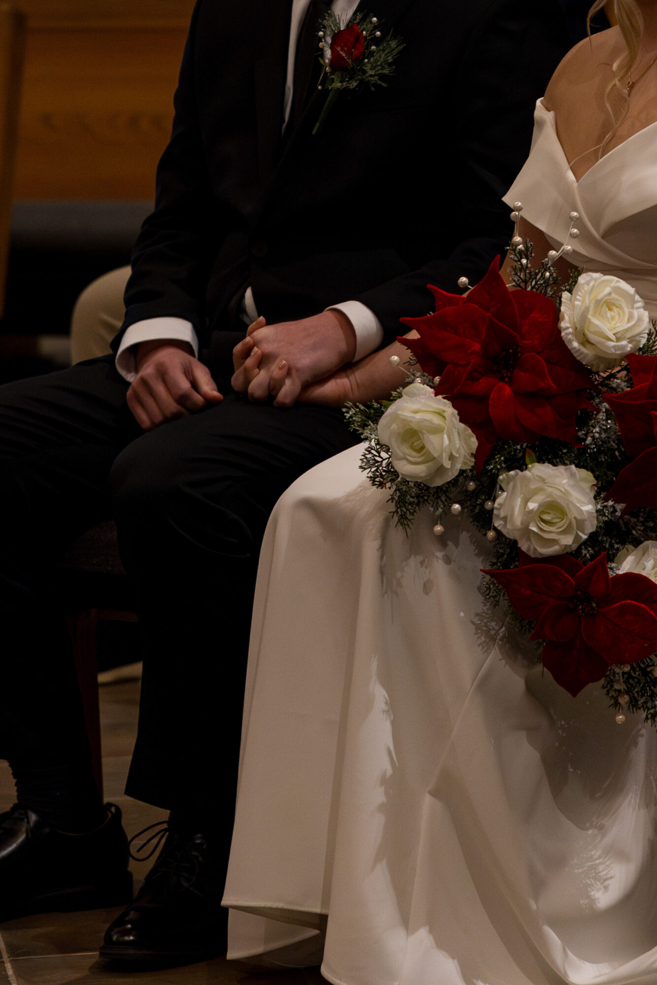 Bride and groom seated, holding hands, with a bouquet of red and white flowers on the bride's lap.