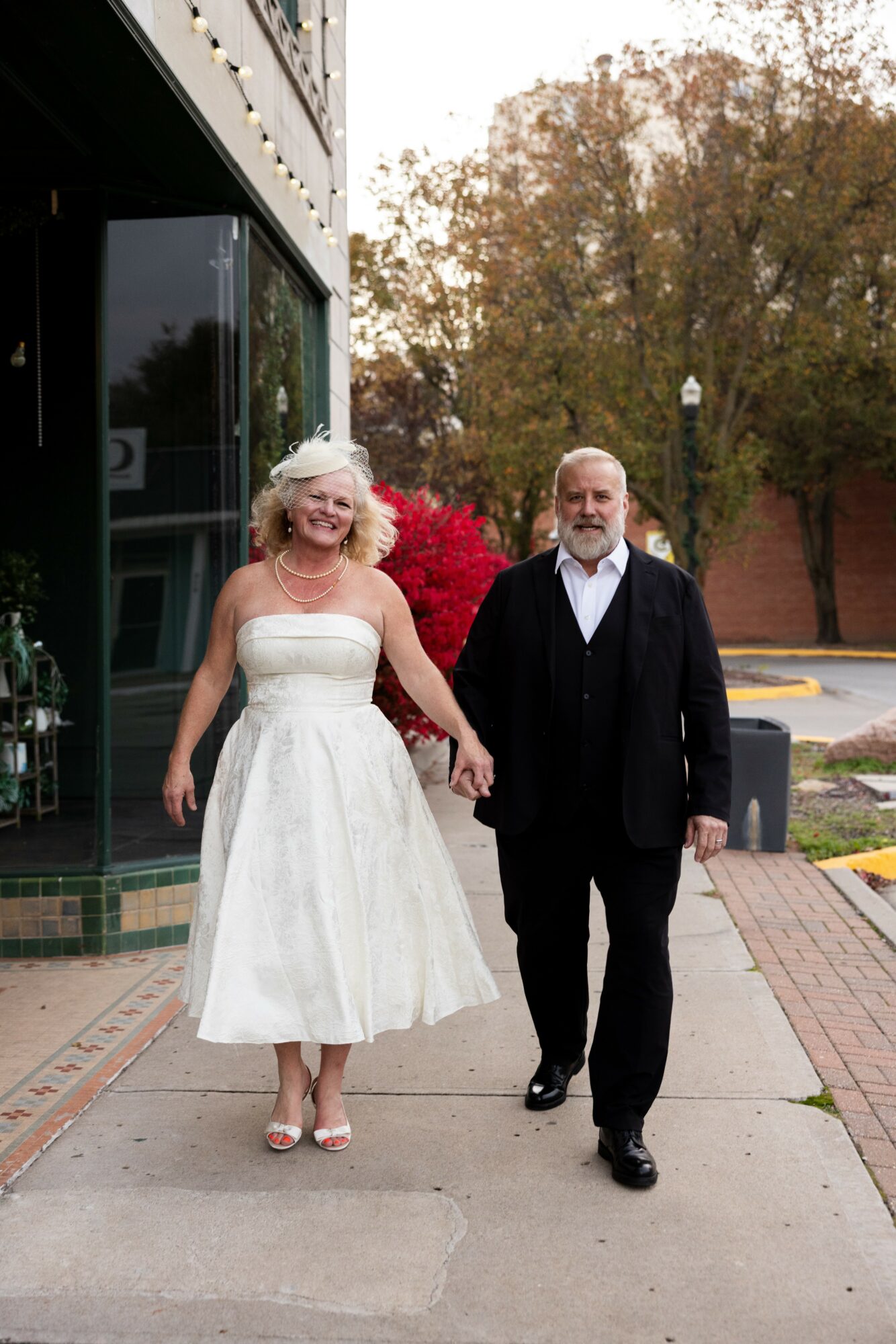 A smiling couple holding hands walking on sidewalk outside building with trees in background.