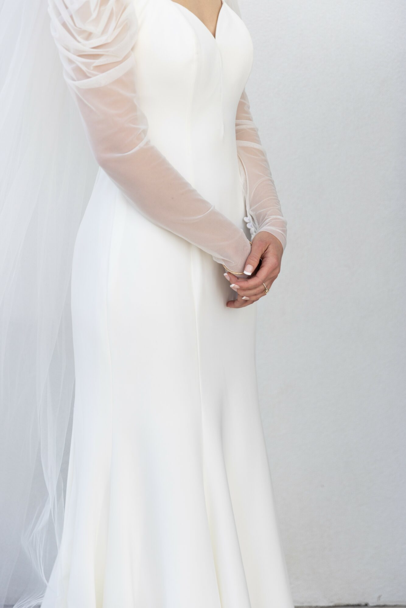 Person in white dress with sheer long sleeves, standing against a plain background, hands clasped in front.