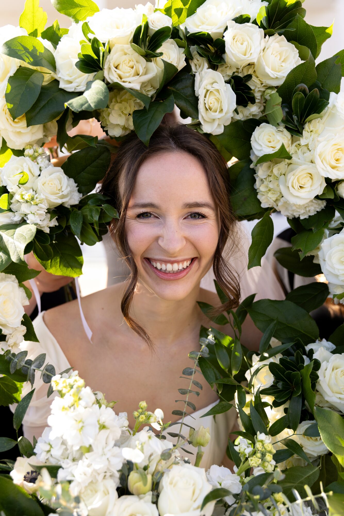 Young woman smiling surrounded by white roses and green leaves, with dark hair and light skin.