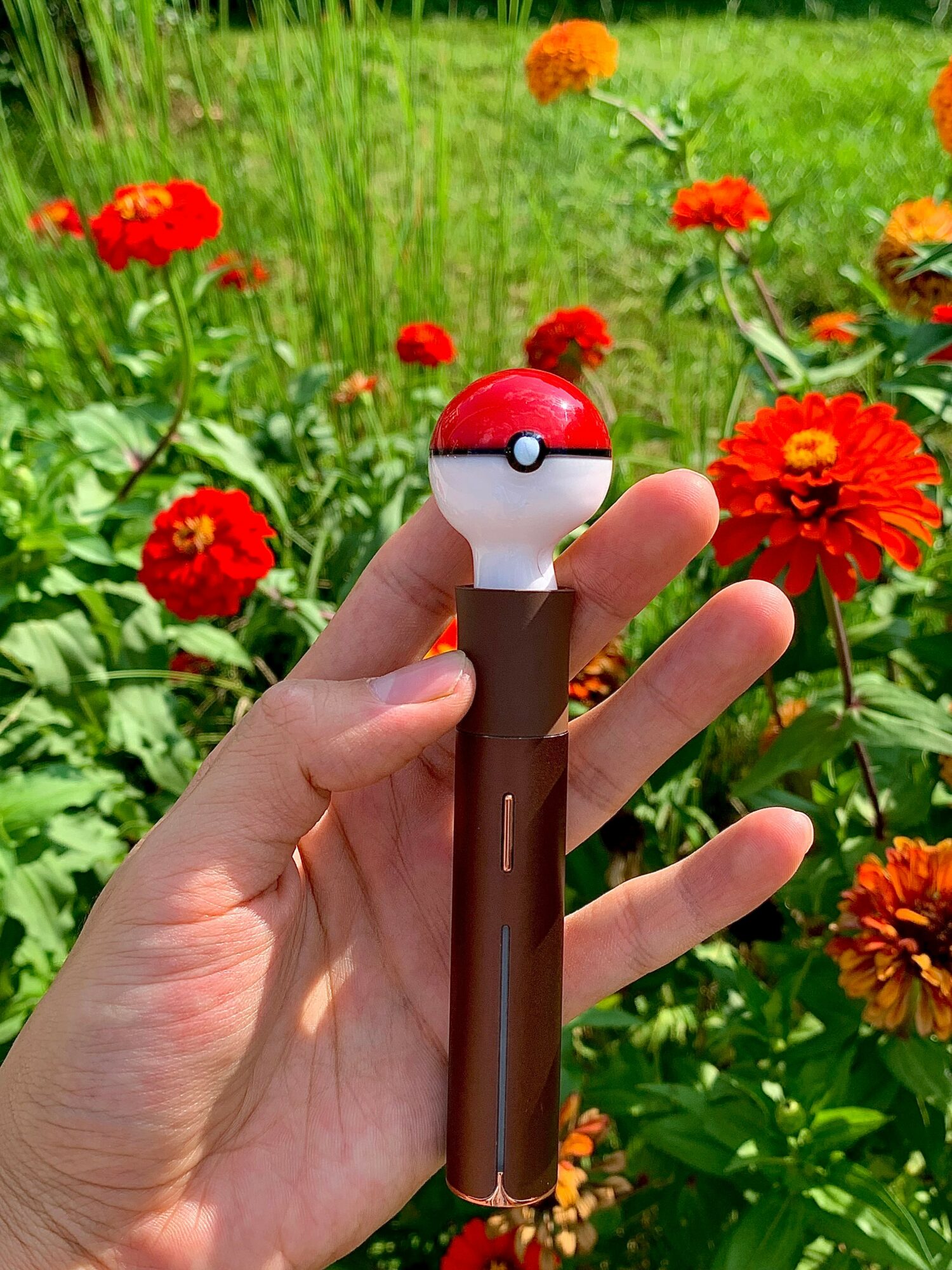 Hand holding a Poké Ball-shaped device with a brown handle, surrounded by colorful flowers in a garden.