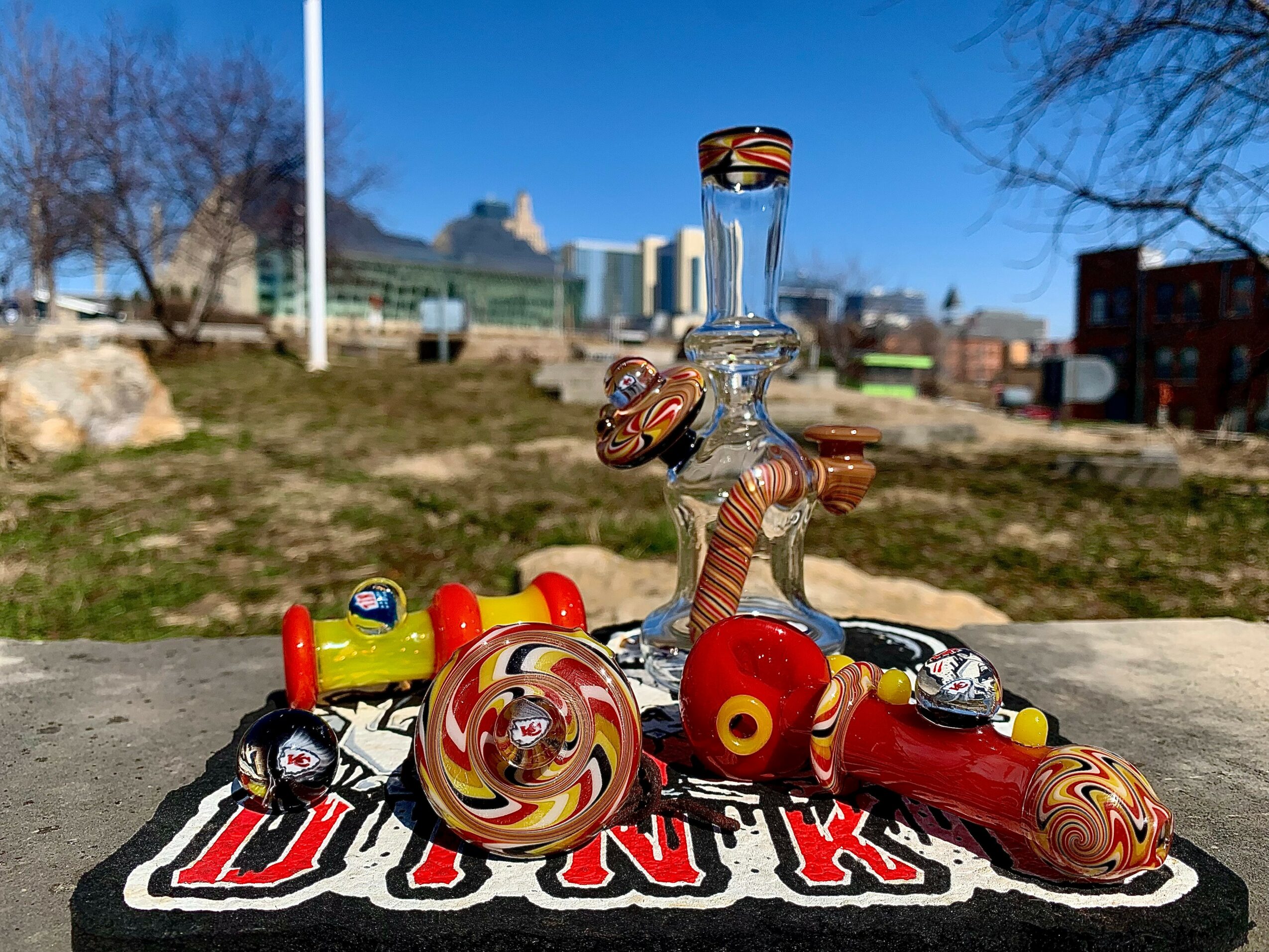 Glass pipe with colorful swirls and a red and yellow ball, placed on a black and red cloth outdoors with trees and buildings in background.