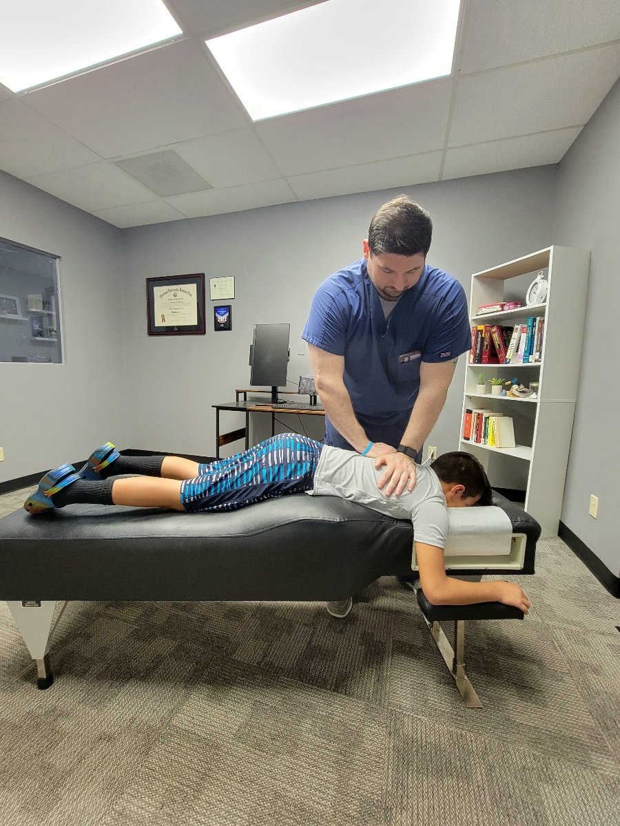Person receiving a back massage from a therapist in an office room with bookshelf and framed certificates.