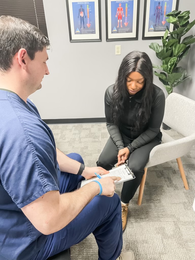 A woman sits in a chair with her head bowed, a man in scrubs holds a clipboard, in a medical office.