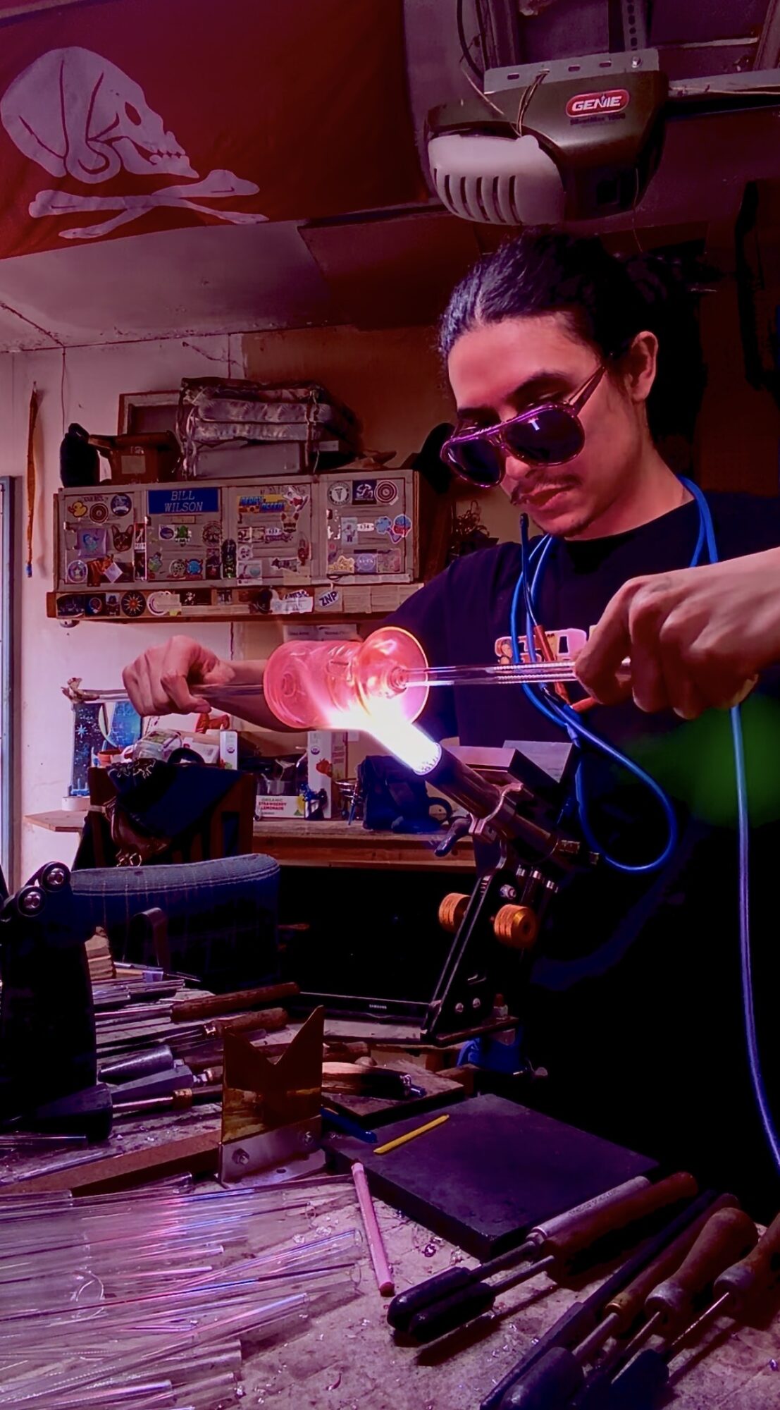Person wearing sunglasses working with glowing glass in a workshop with tools and equipment.