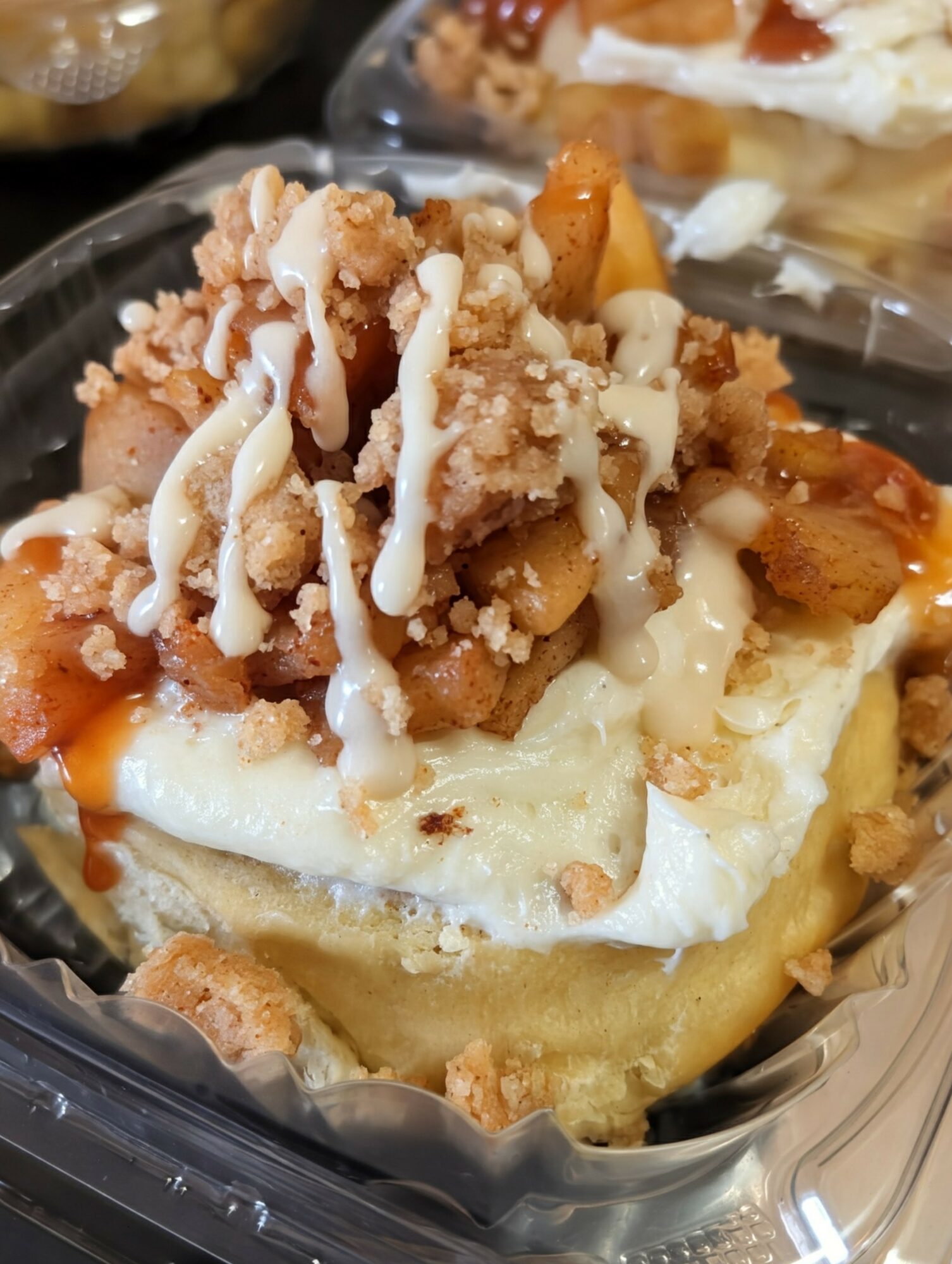 Close-up of a dessert with crumb topping, white drizzle, and a creamy base, in a clear container.