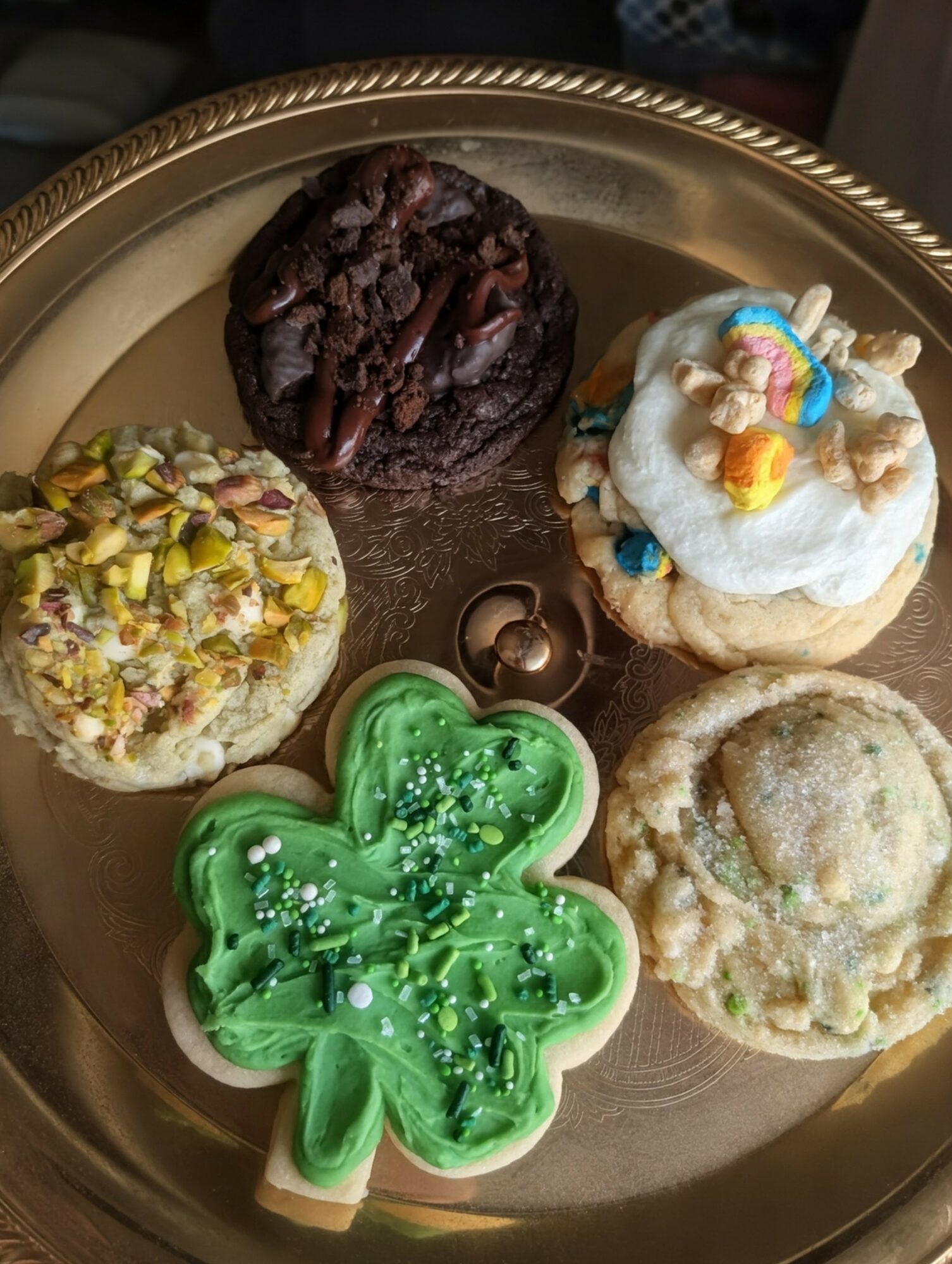 Plate with four decorated cookies, including a green shamrock, a white cookie with colorful candies, a chocolate cookie, and a cookie with crushed nuts.