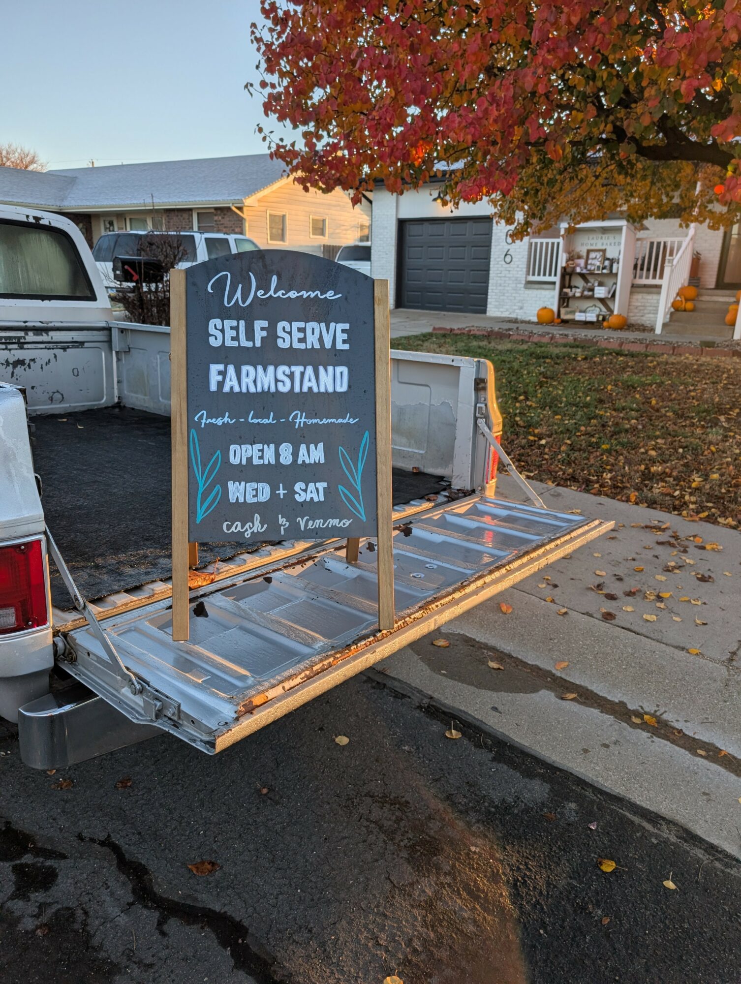 Chalkboard sign on a truck bed advertising self-serve farmstand open Wednesday to Saturday, with fall leaves and a tree in background.