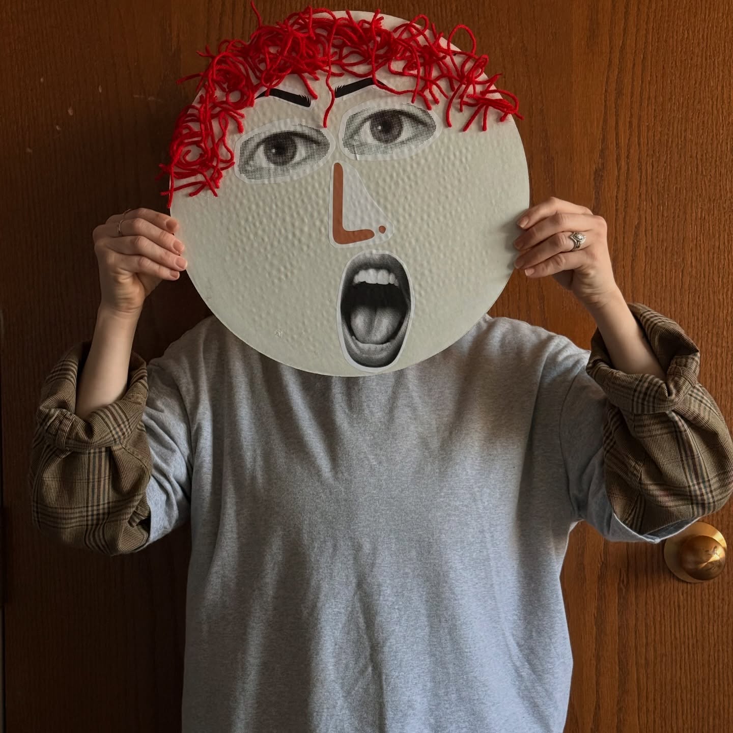 Person holding a large, round, white mask with eyes, nose, mouth, and red curly hair, covering their face.