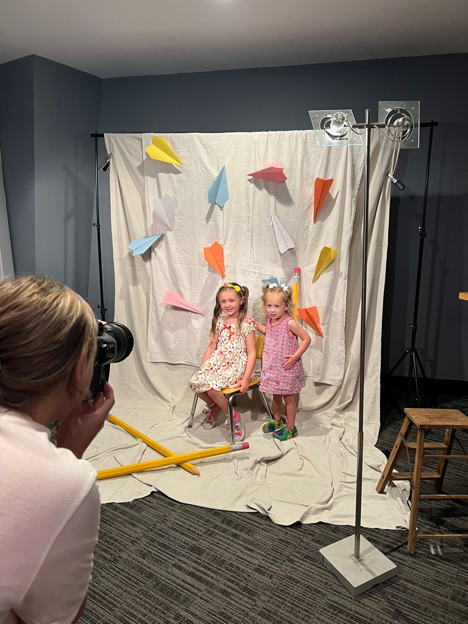 Two young girls sit on a white fabric backdrop with colorful paper hats, posing for a photo in a room with blue walls.
