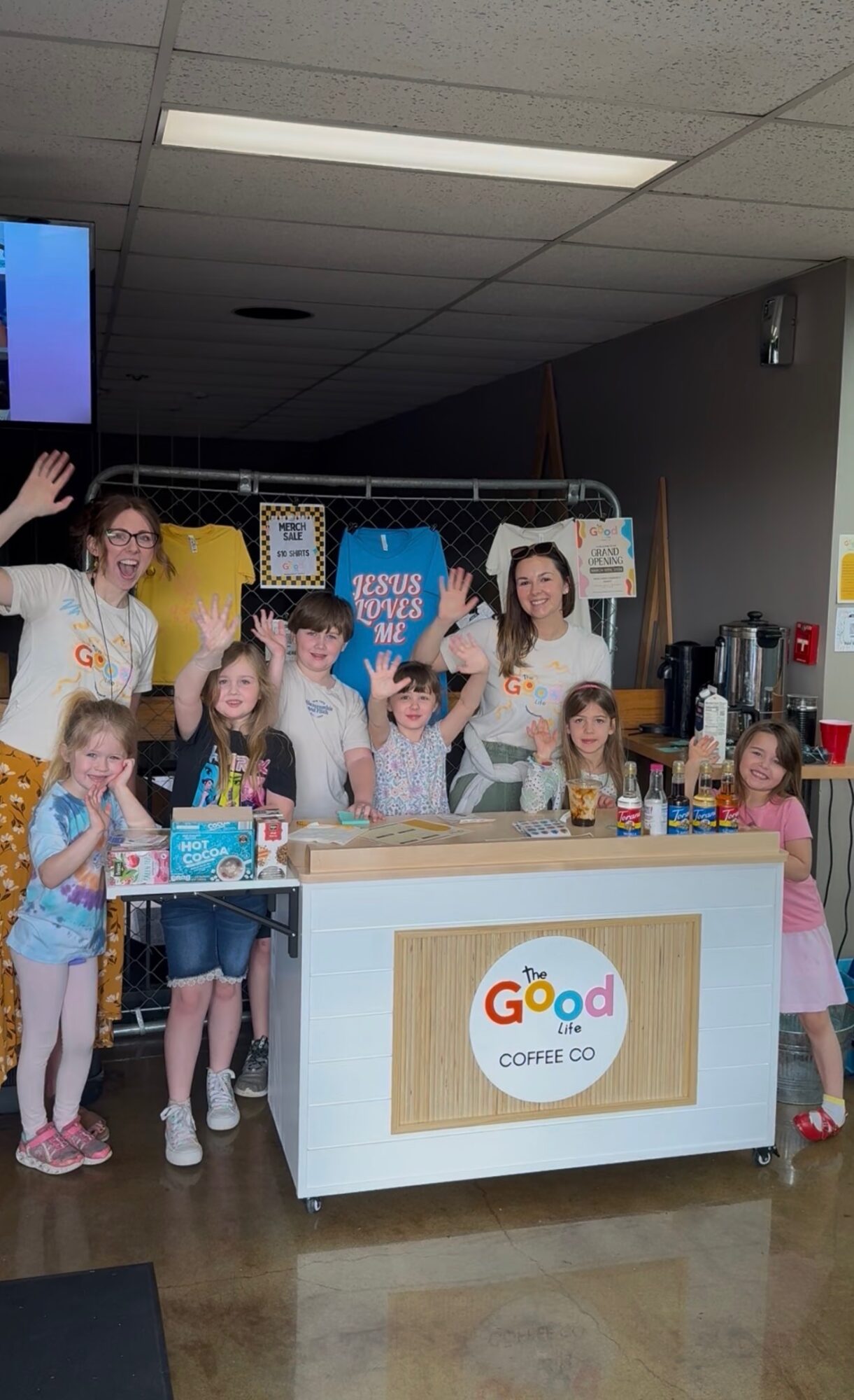 Group of children and adults behind a coffee stand with a colorful sign, in an indoor setting.