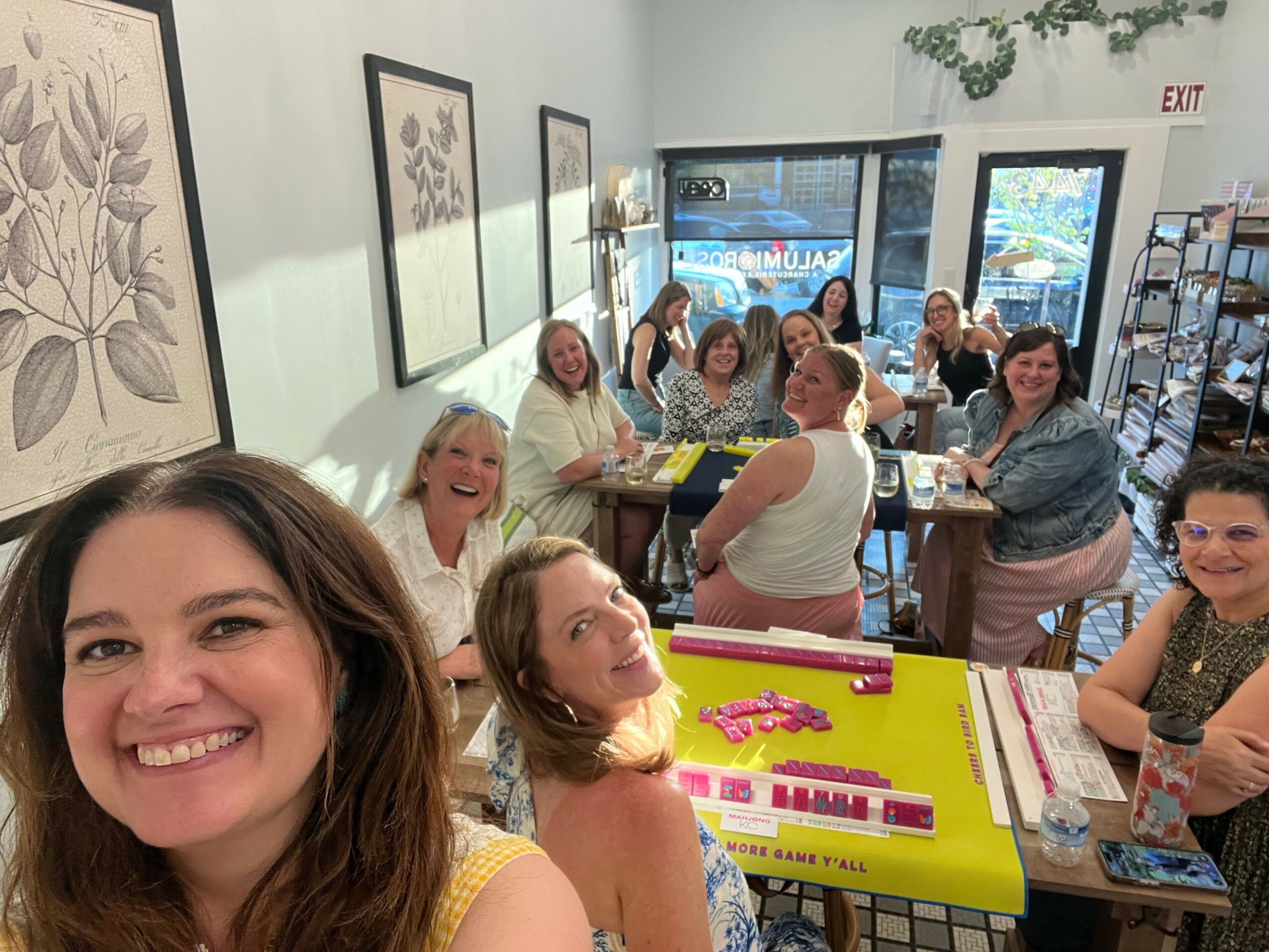 Group of women sitting around tables in a cafe, smiling and chatting, with artwork on the wall behind them.