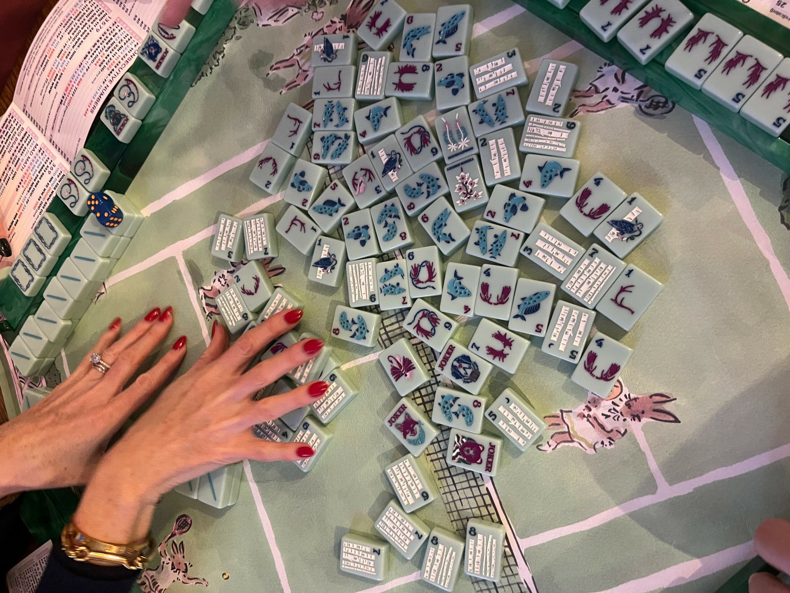 Hands with red nail polish and a gold watch reach towards small boxes with colorful illustrations on a table.