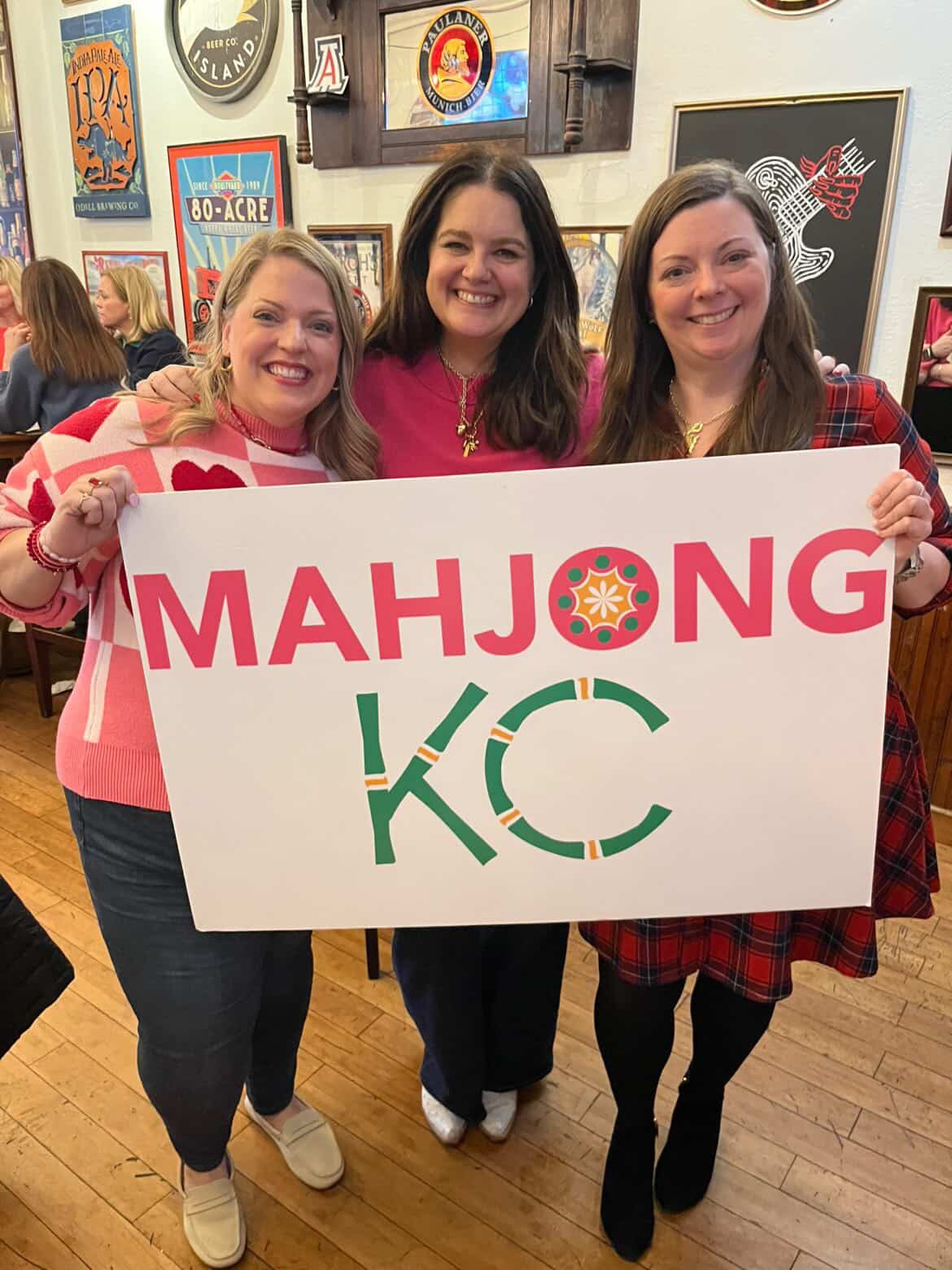 Three women smiling and holding a sign that says 'MAHJONG KC' in a decorated indoor space.