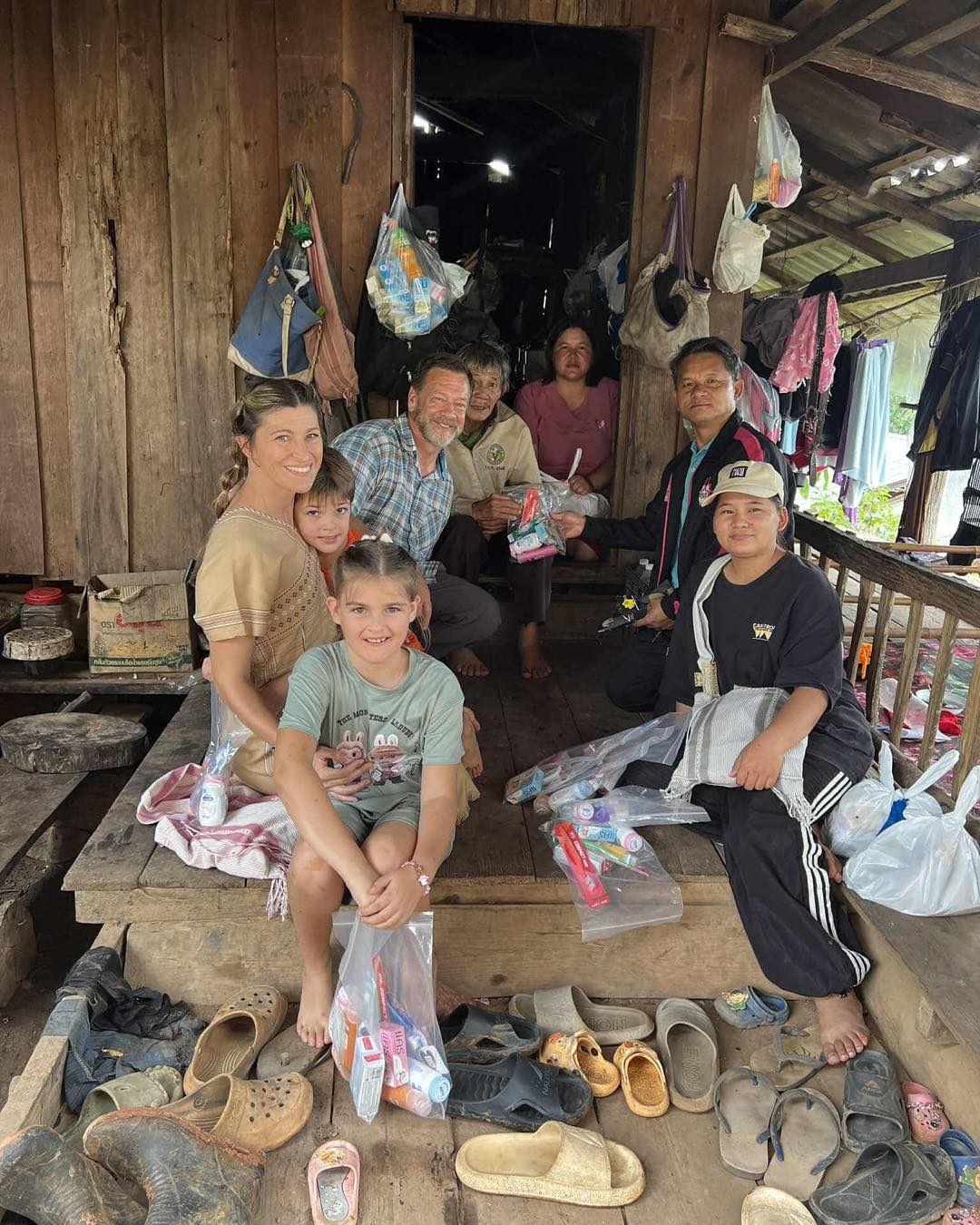 Group of children and adults inside a wooden structure with hanging bags, sitting and standing around shoes and packages.