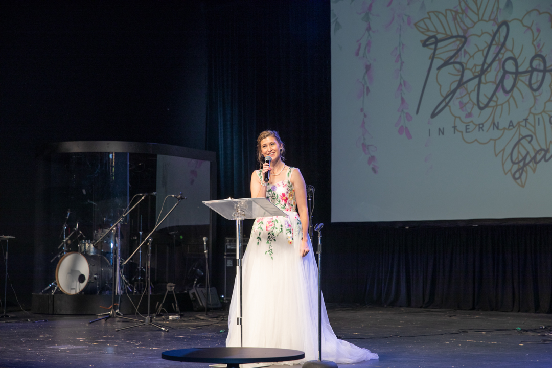 Woman in a white dress speaking at a podium on stage with a large screen behind her.