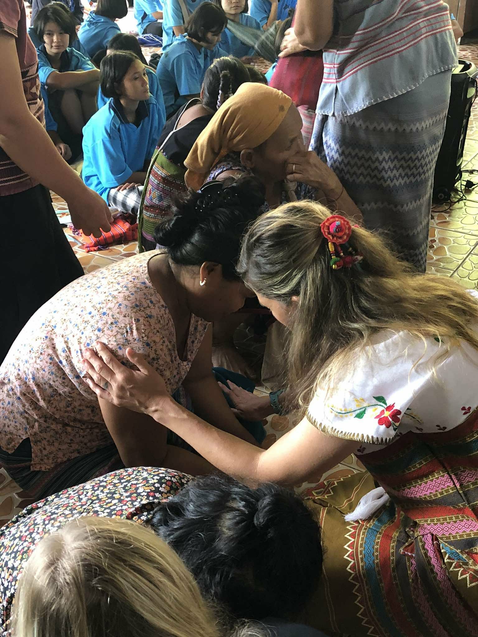 Two women engage in a close interaction, surrounded by children and adults in a crowded indoor setting.