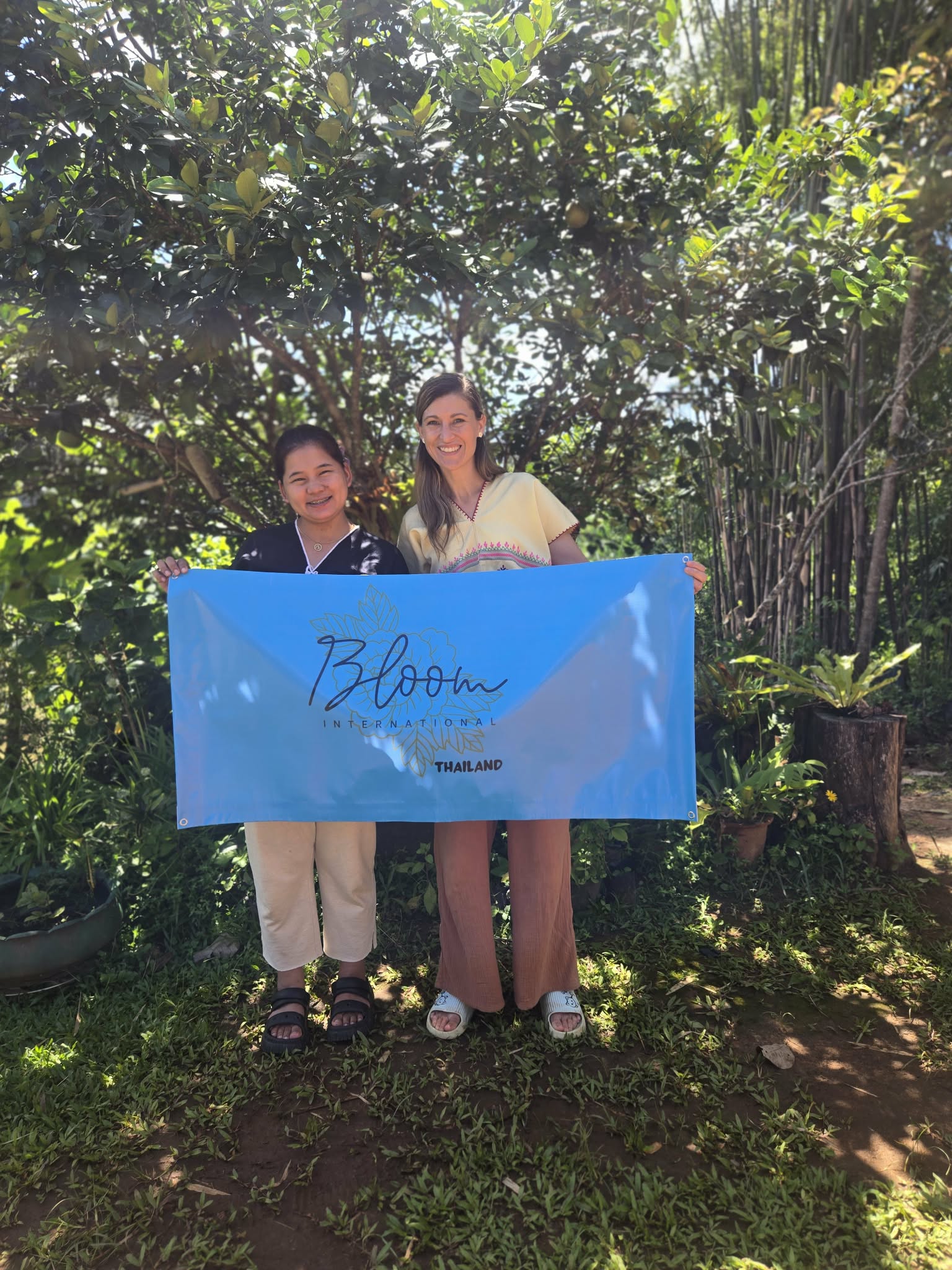Two women standing outdoors holding a blue banner with the word 'Bloom' in a garden setting.