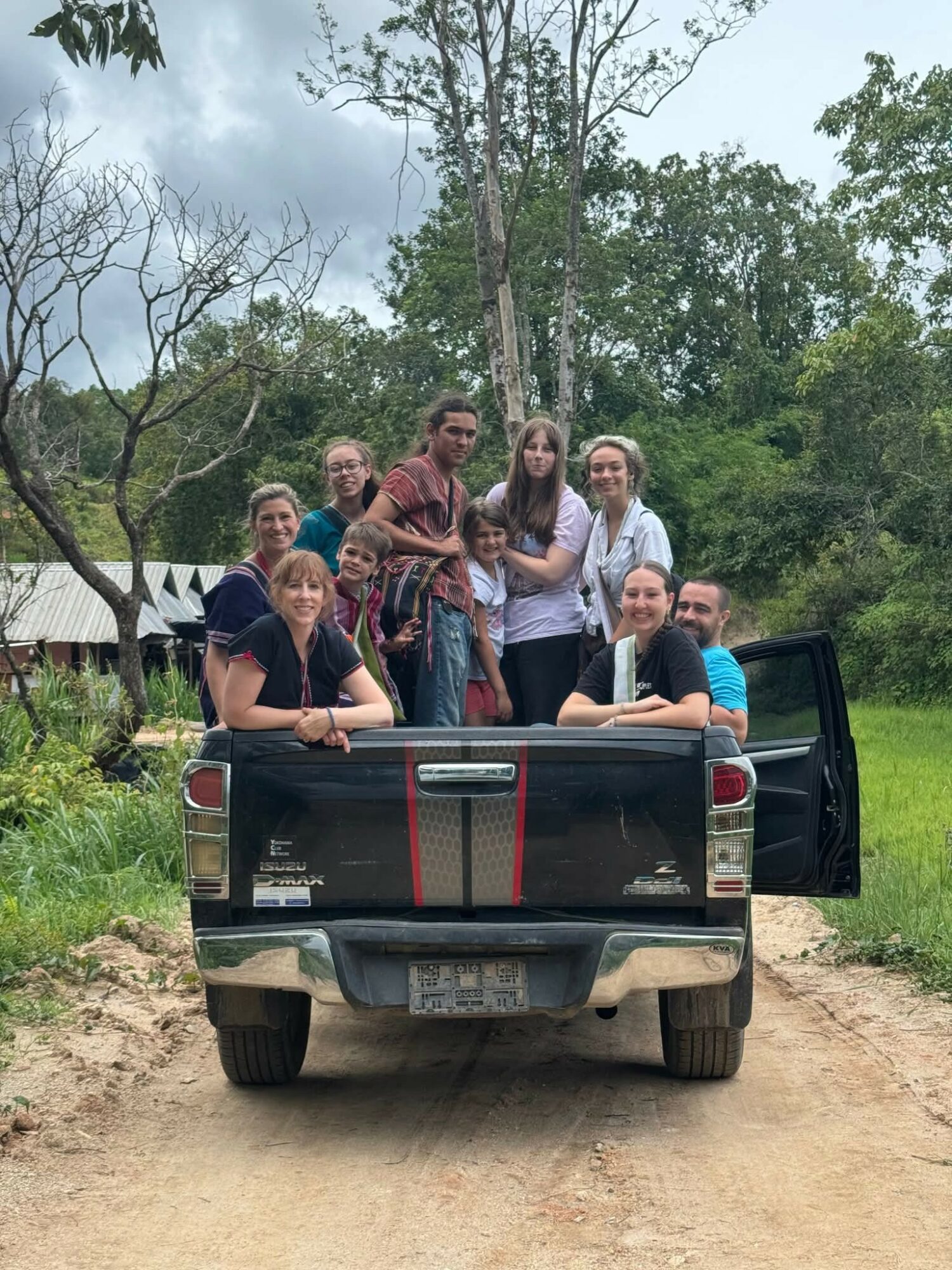 Group of people sitting and standing in the back of a black pickup truck on a dirt road, surrounded by trees and greenery.
