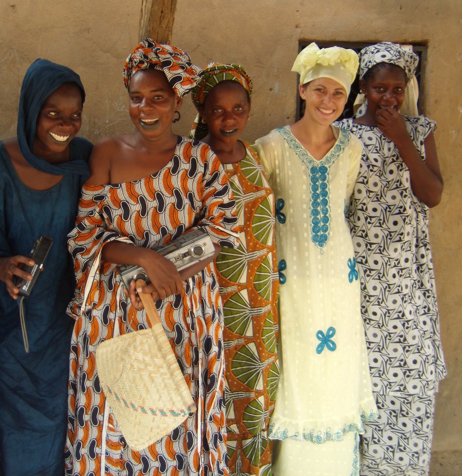 Five women standing together, smiling, wearing colorful traditional and modern clothing, in front of a plain wall.