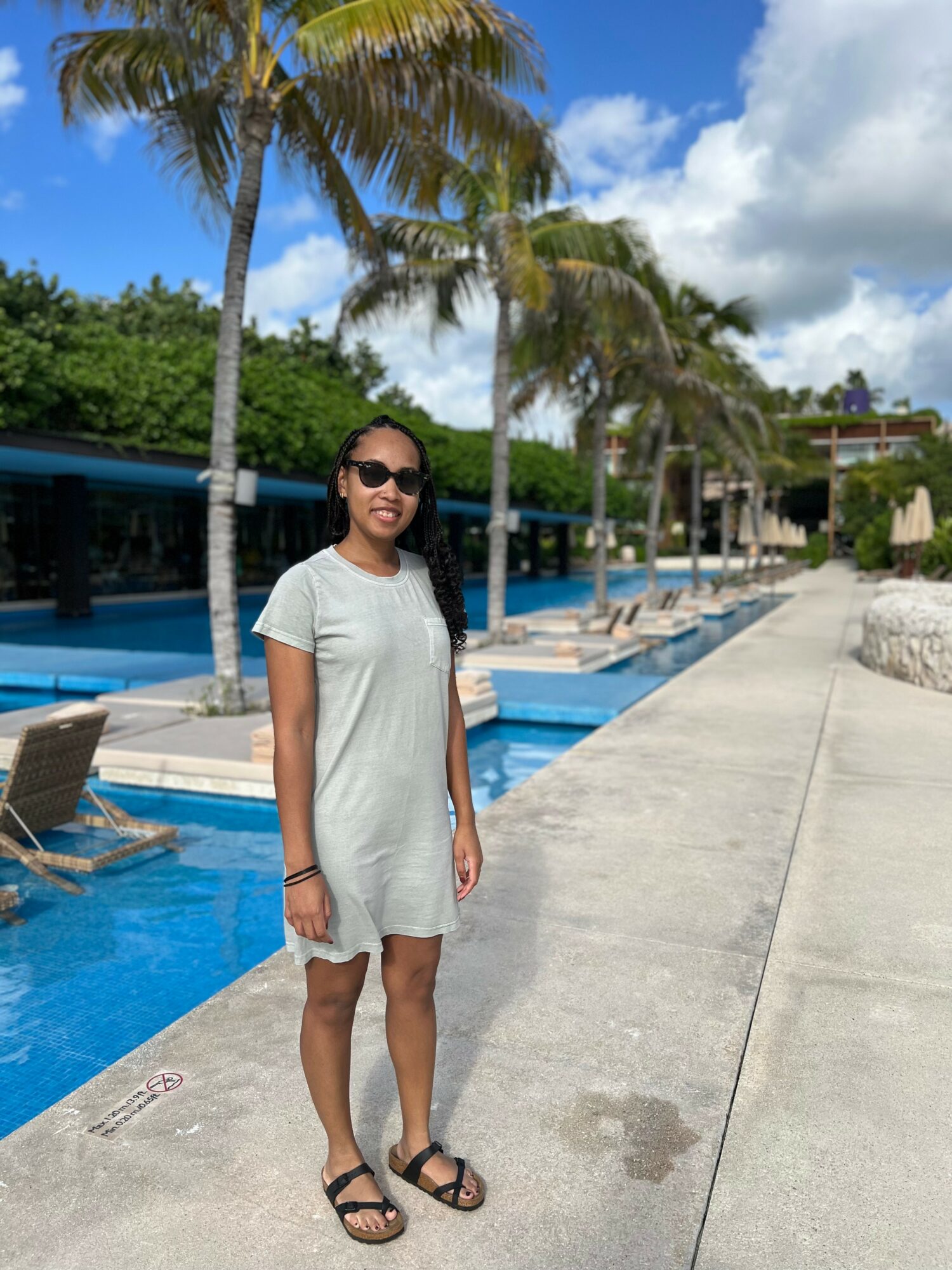 Woman in sunglasses and casual dress standing near poolside with palm trees and lounge chairs in background.