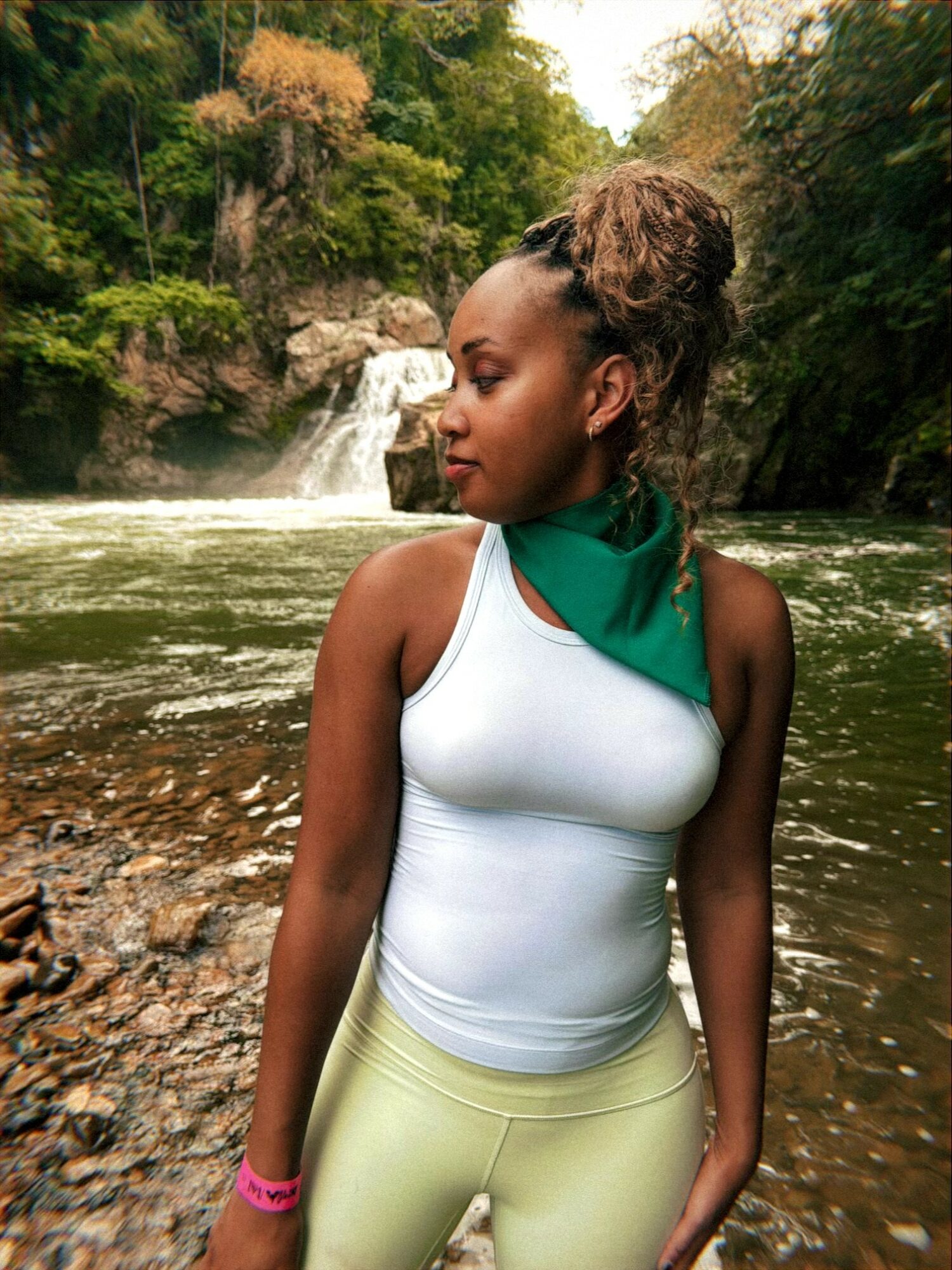 Young woman with braided hair in a bun, wearing a white tank top and green scarf, standing by a river with waterfall.