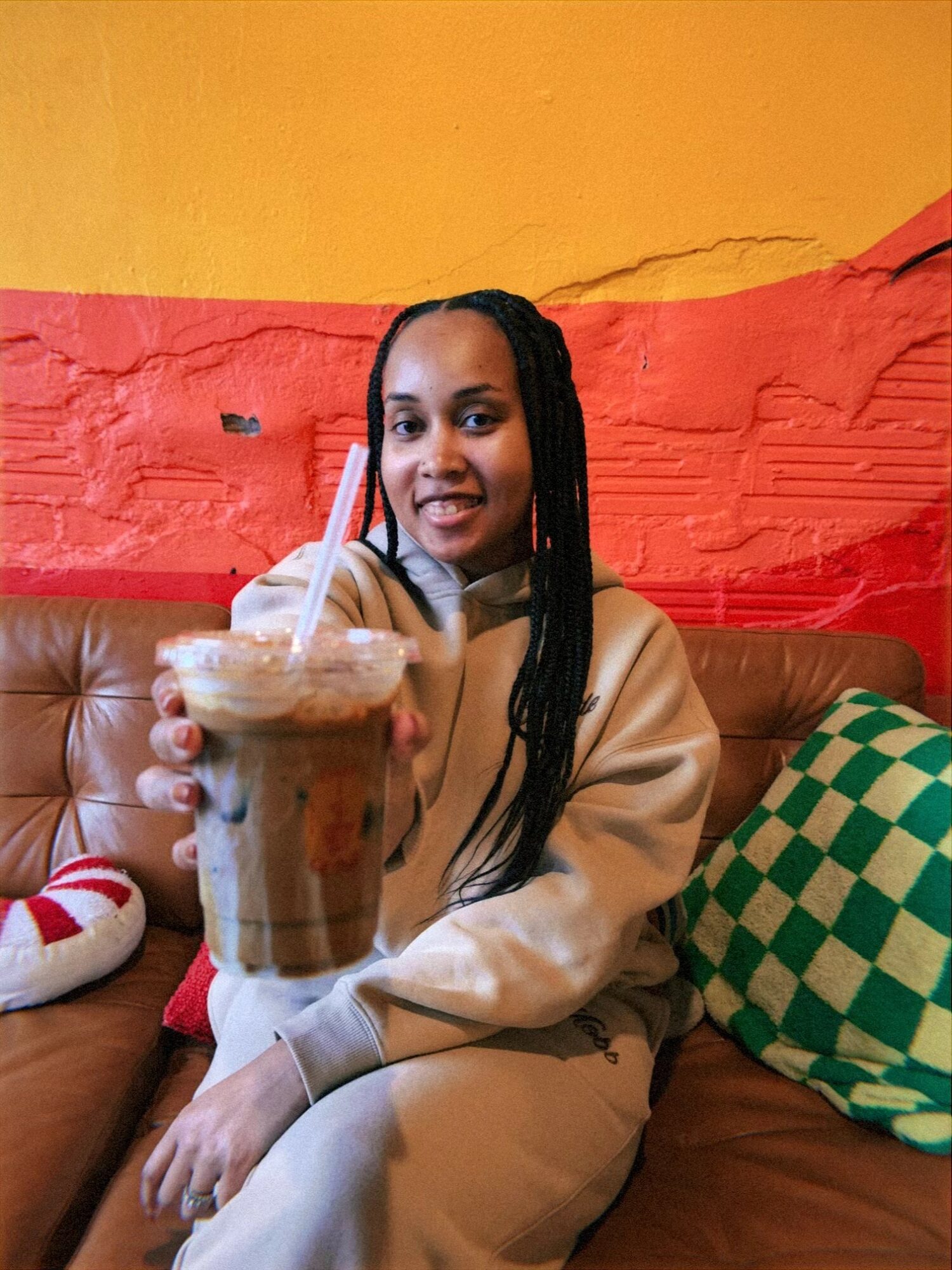 Woman sitting on a couch holding a large iced coffee with a straw, smiling at the camera.