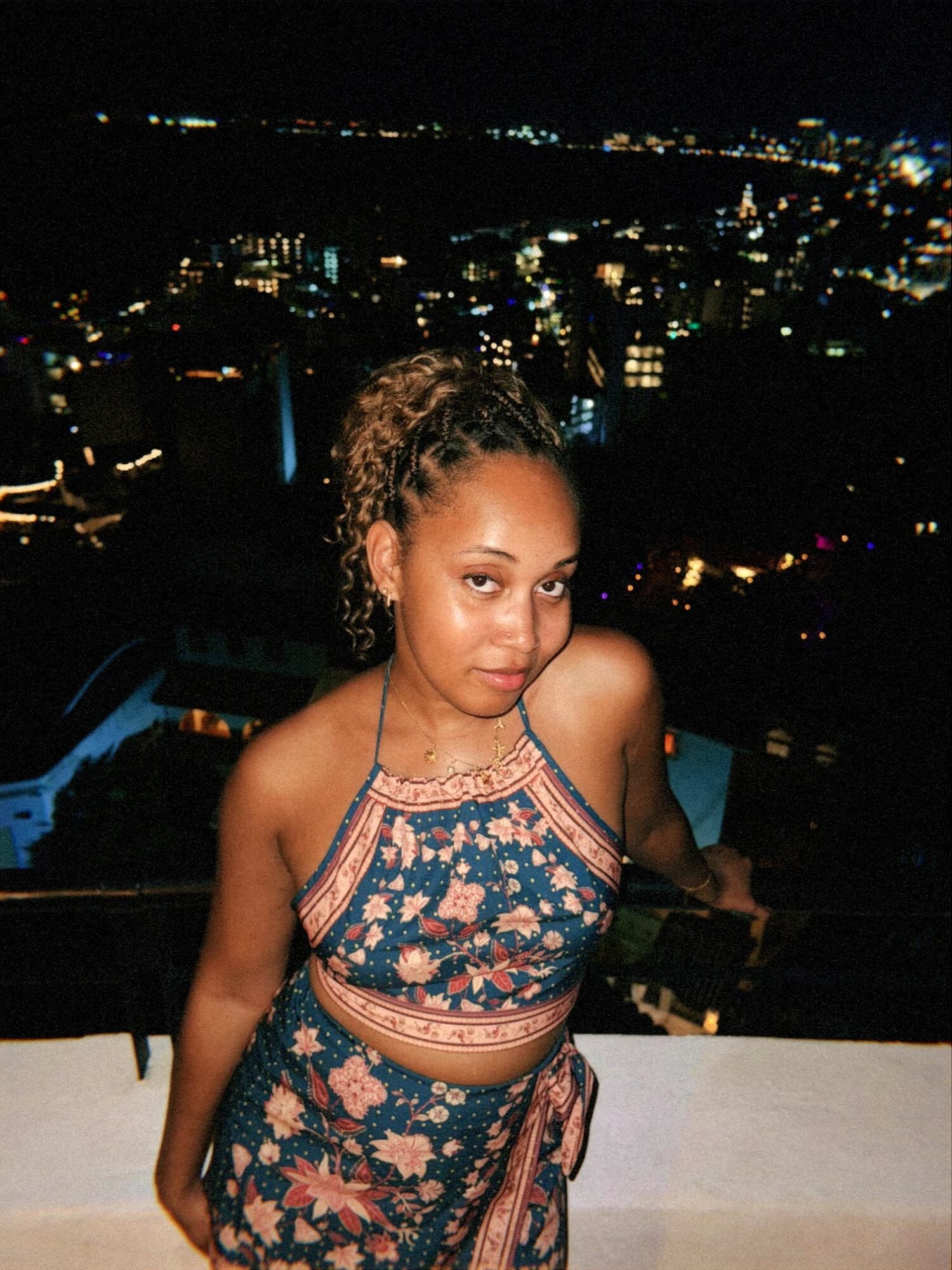 Young woman with curly hair in a patterned dress standing on a balcony at night, city lights in background.