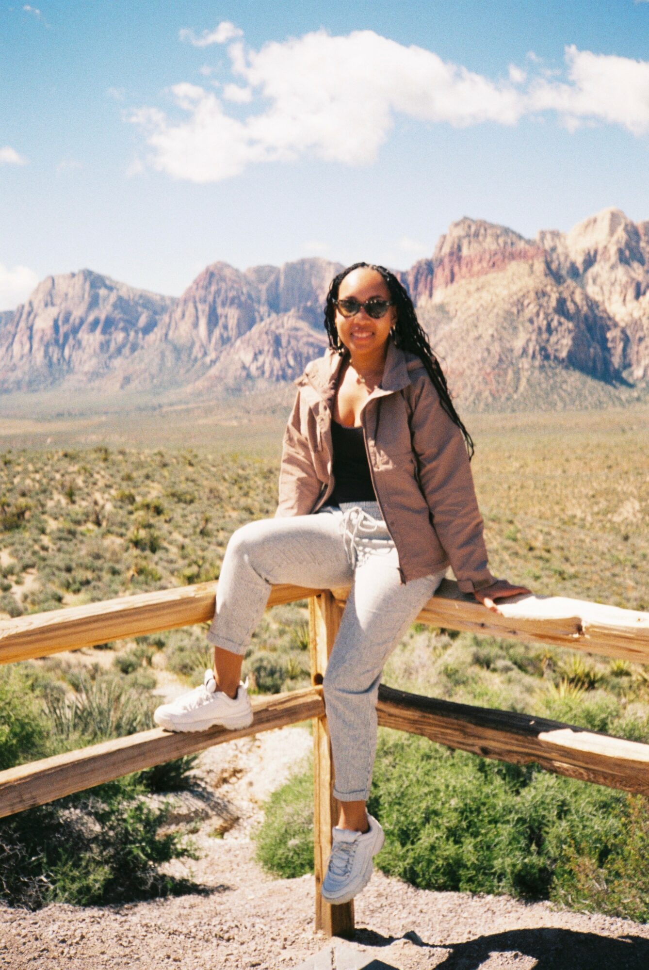 Woman sitting on a wooden fence in a desert landscape with mountains and blue sky.