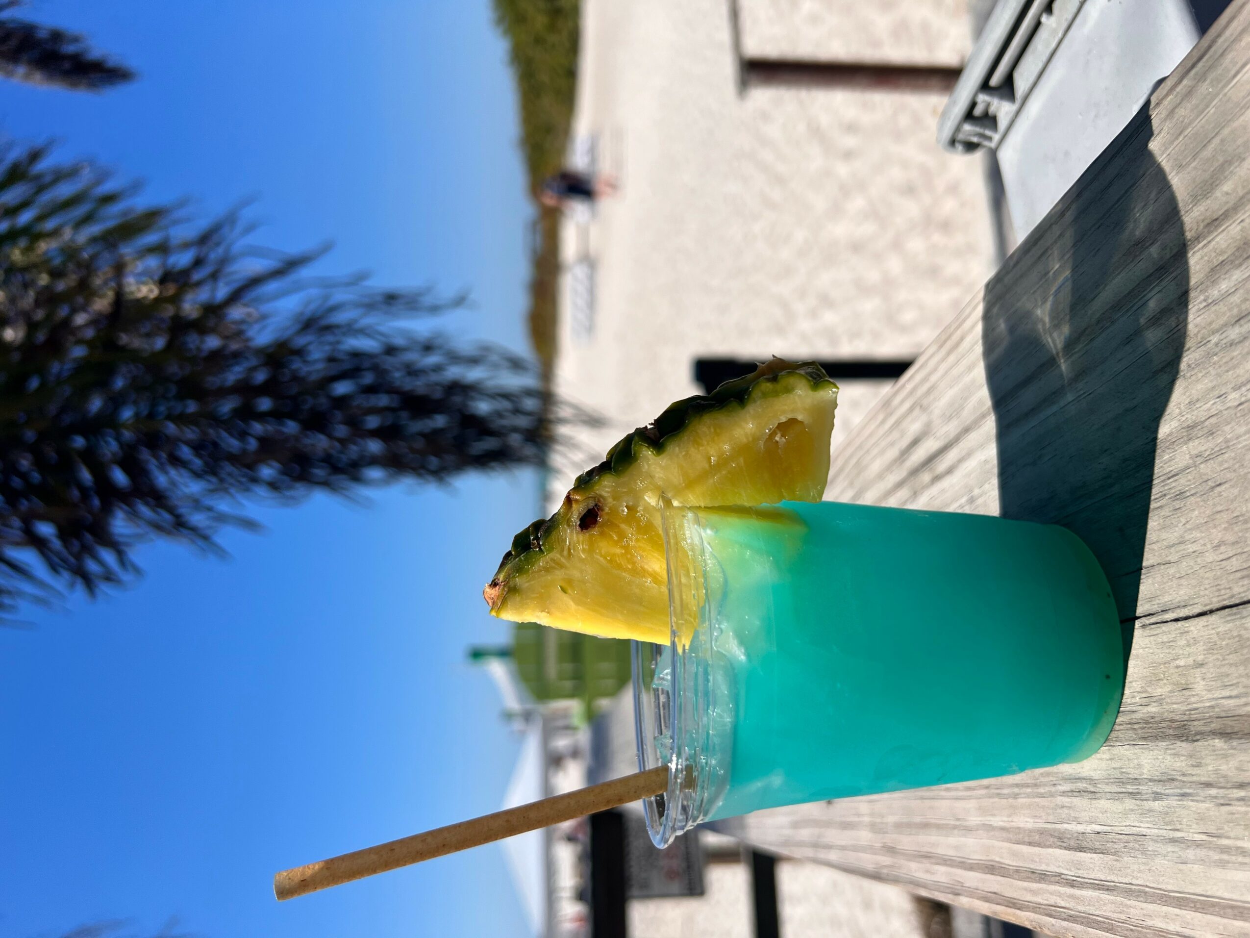 Blue drink with pineapple slice and straw on wooden surface outdoors, with trees and buildings in background.