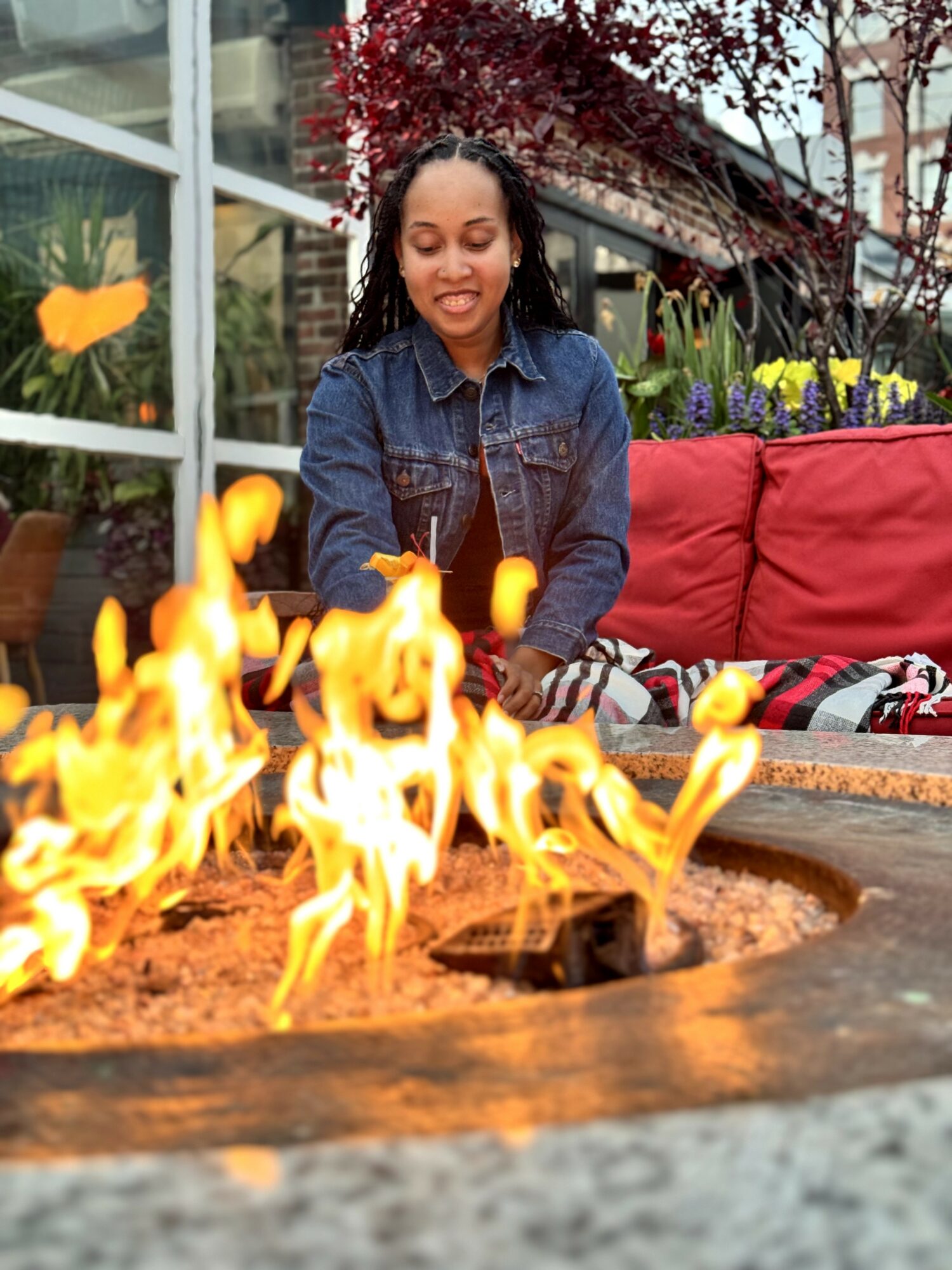 Young woman with braided hair in denim jacket sitting outdoors near a fire pit with flames.