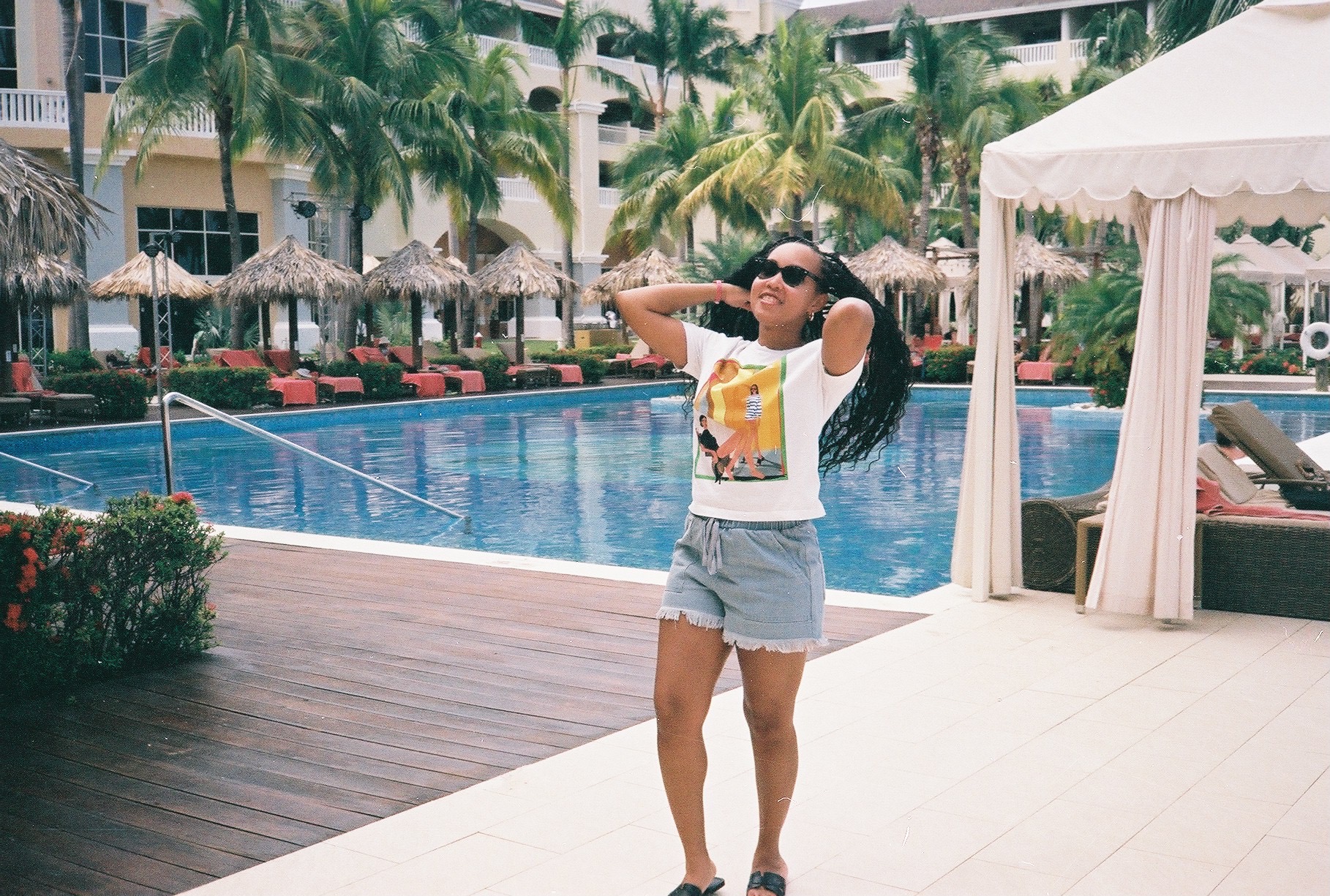 Woman in sunglasses and casual clothes standing near poolside with arms outstretched, palm trees and umbrellas in background.