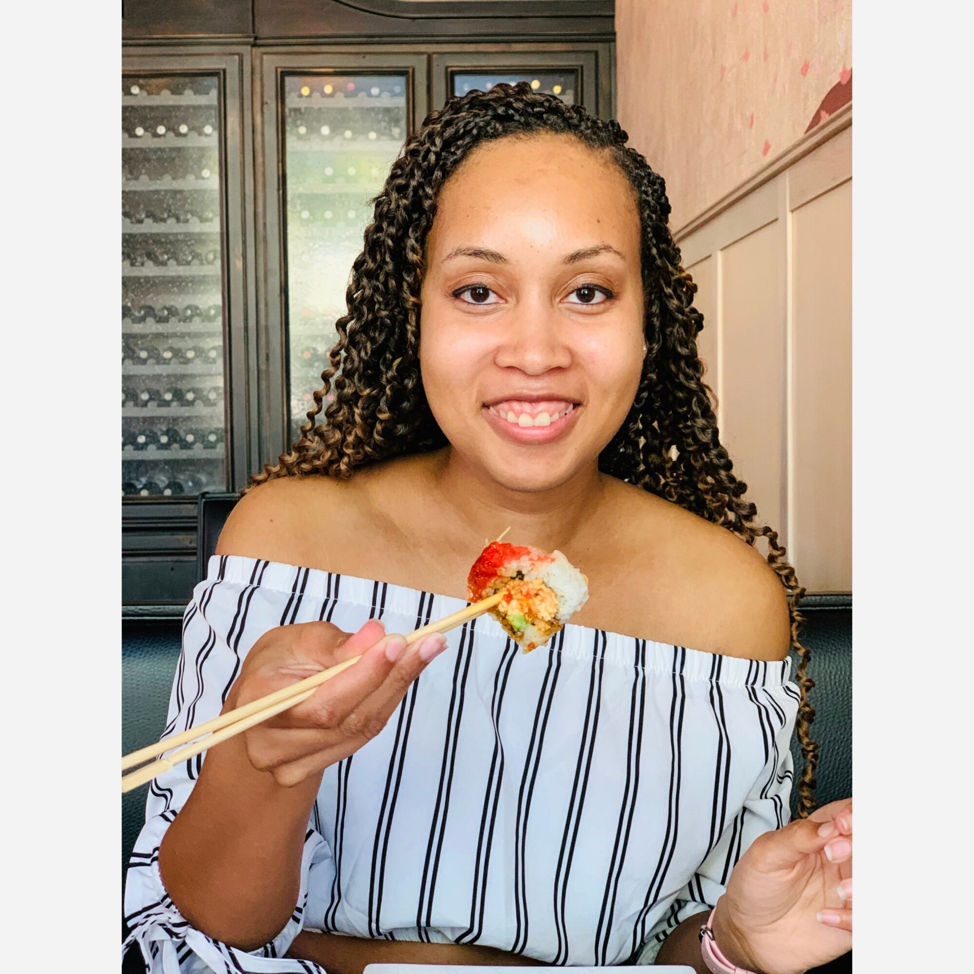 Woman with curly hair holding sushi with chopsticks, smiling indoors, wearing an off-shoulder striped top.
