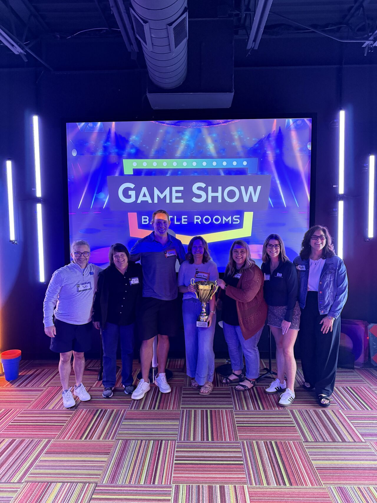 Group of eight people standing in front of a large screen with a trophy, in a game show setting.