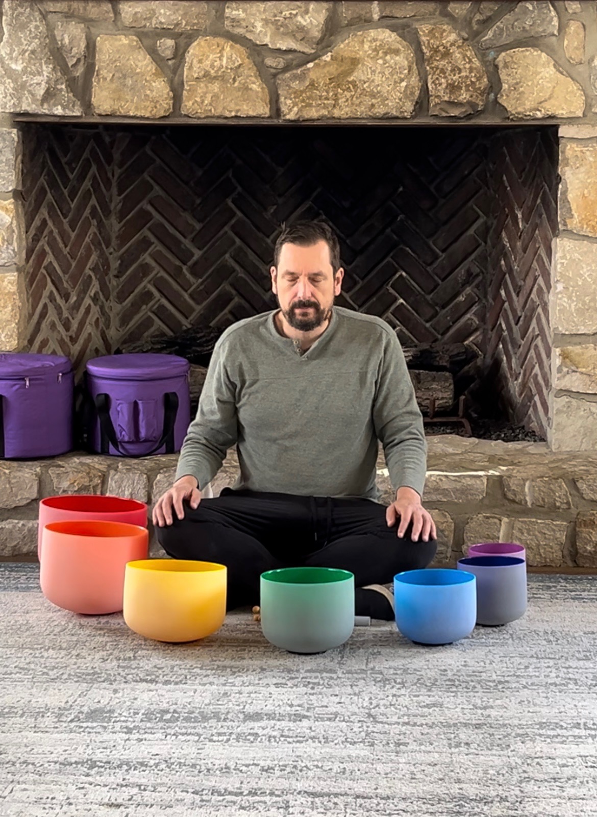 Man sitting cross-legged in front of a fireplace with colorful bowls arranged in front of him.