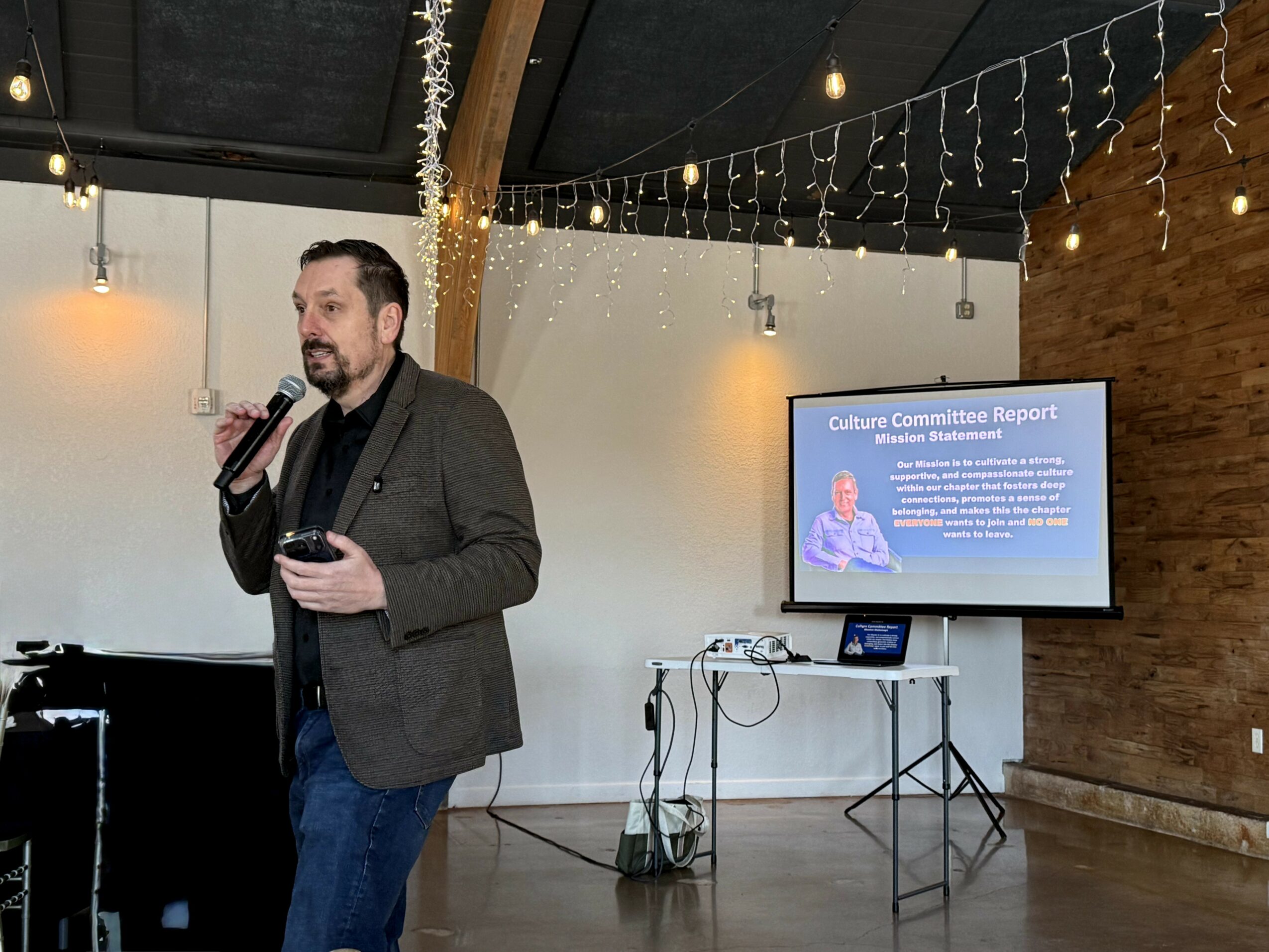 Man in blazer holding microphone and tablet, speaking in a room with string lights and a presentation screen.