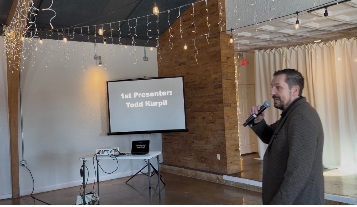 Man speaking into microphone at presentation event with screen displaying name and title.
