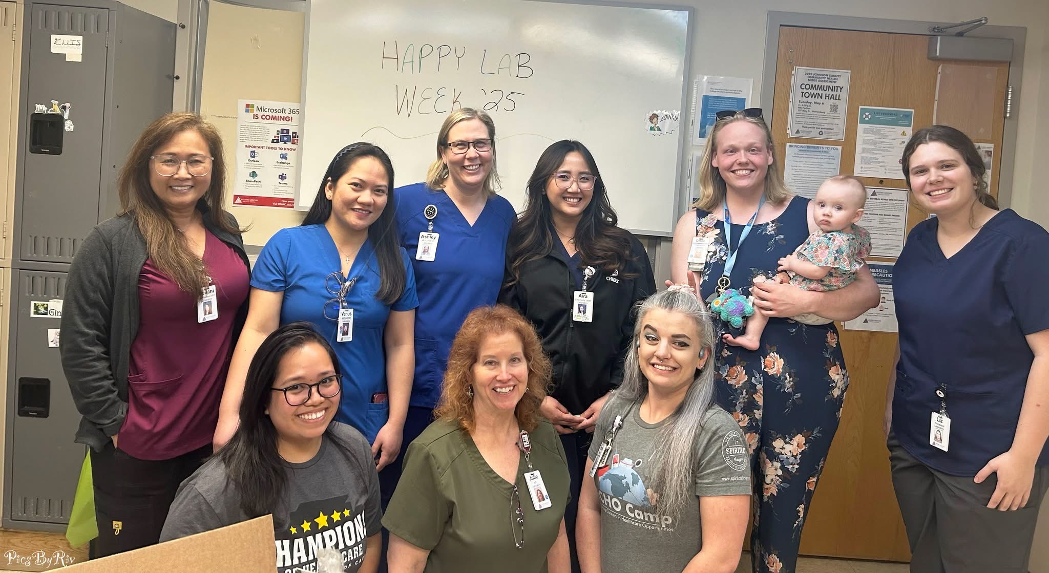Group of nine women and one baby in a classroom, some wearing scrubs and ID badges, smiling at the camera.