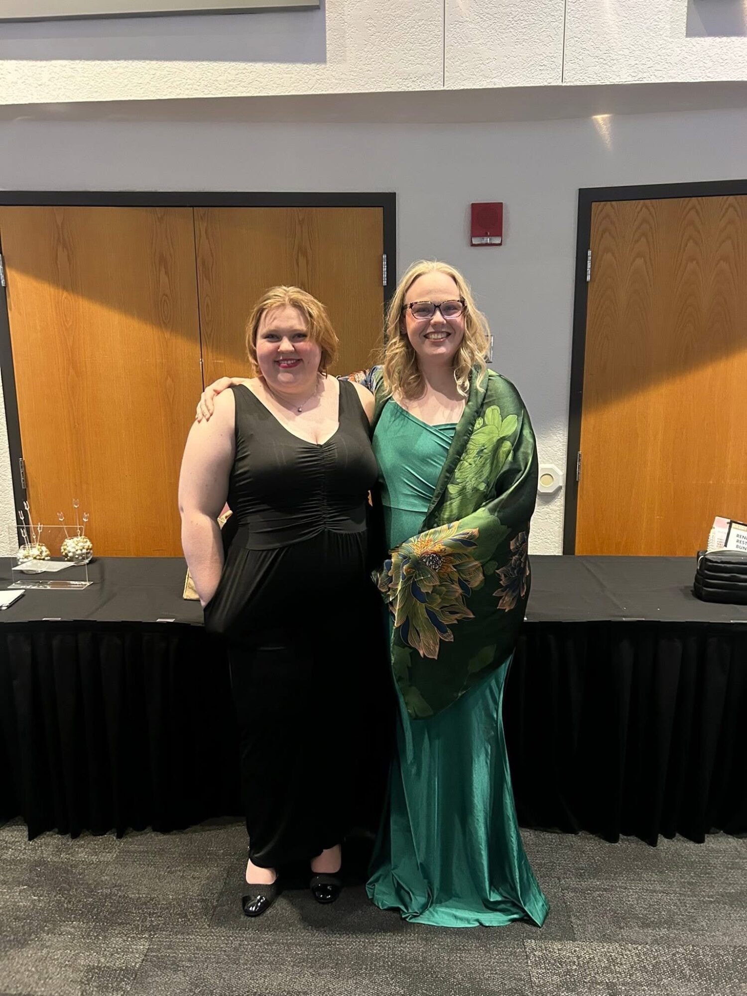 Two women in formal dresses standing side by side indoors, smiling at the camera, with a black table behind them.