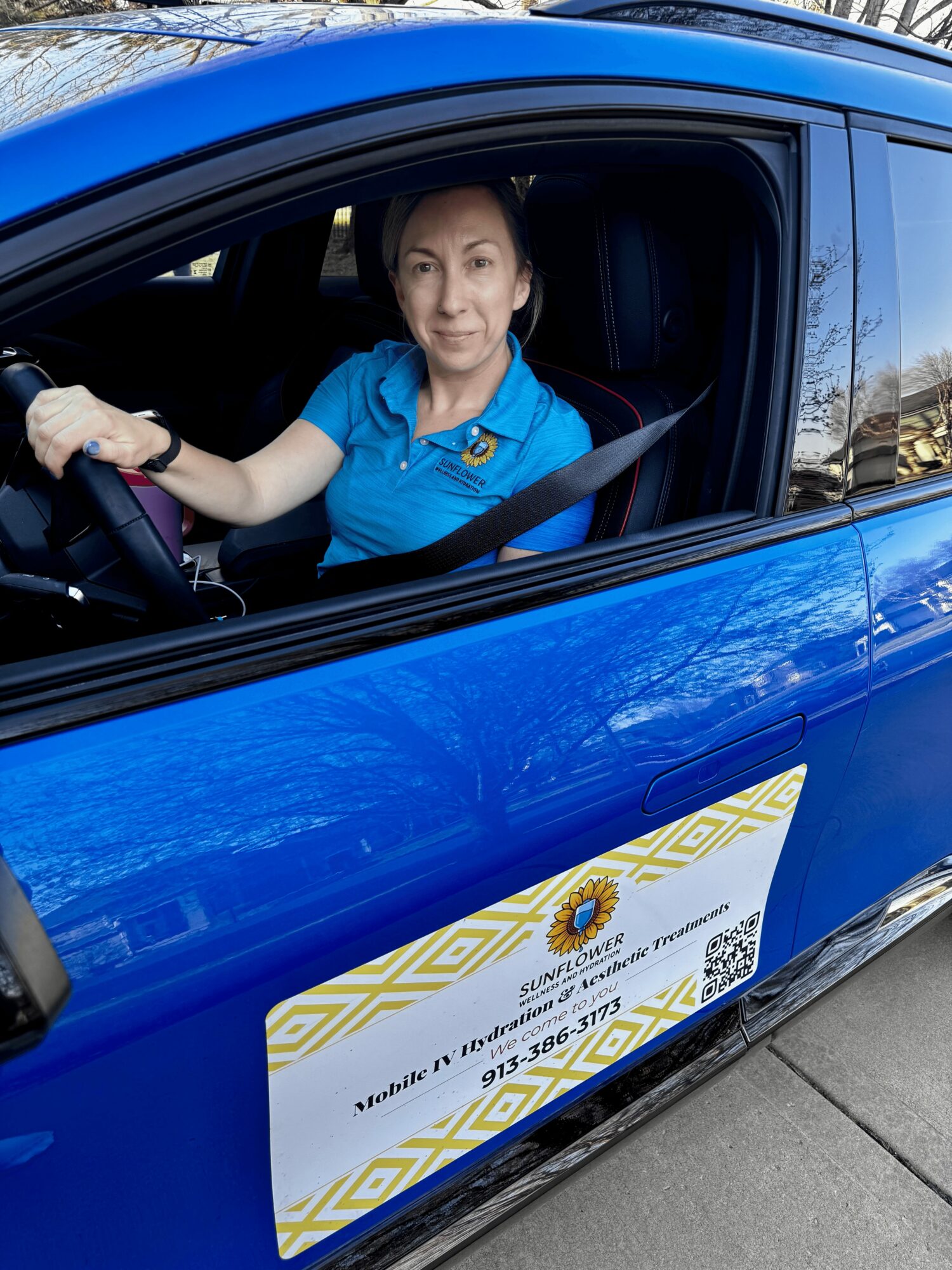 Woman sitting in driver's seat of blue car, looking at camera, with a business card on the door.