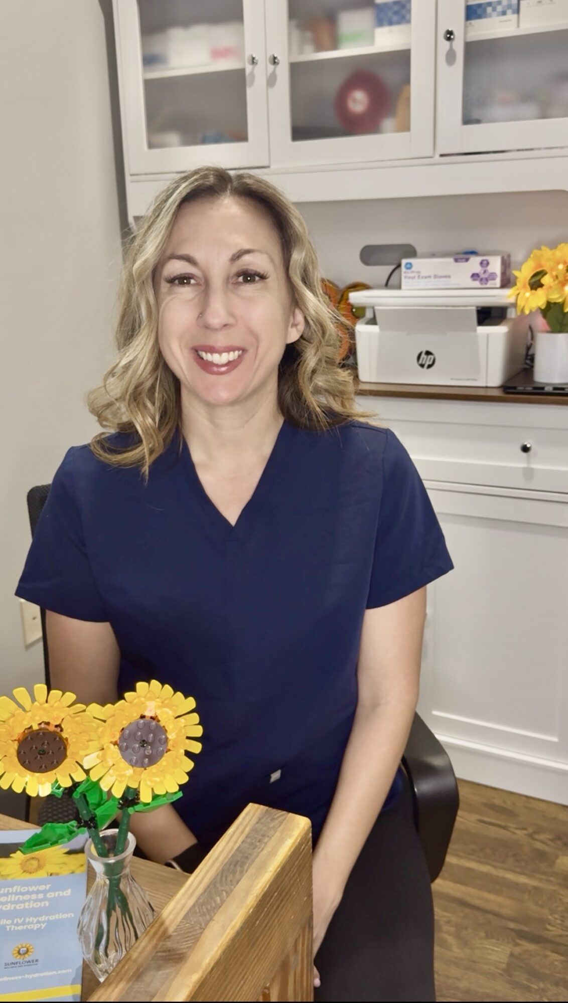 Woman with blonde hair wearing navy scrubs sitting at a table in a room with white cabinets and flowers.