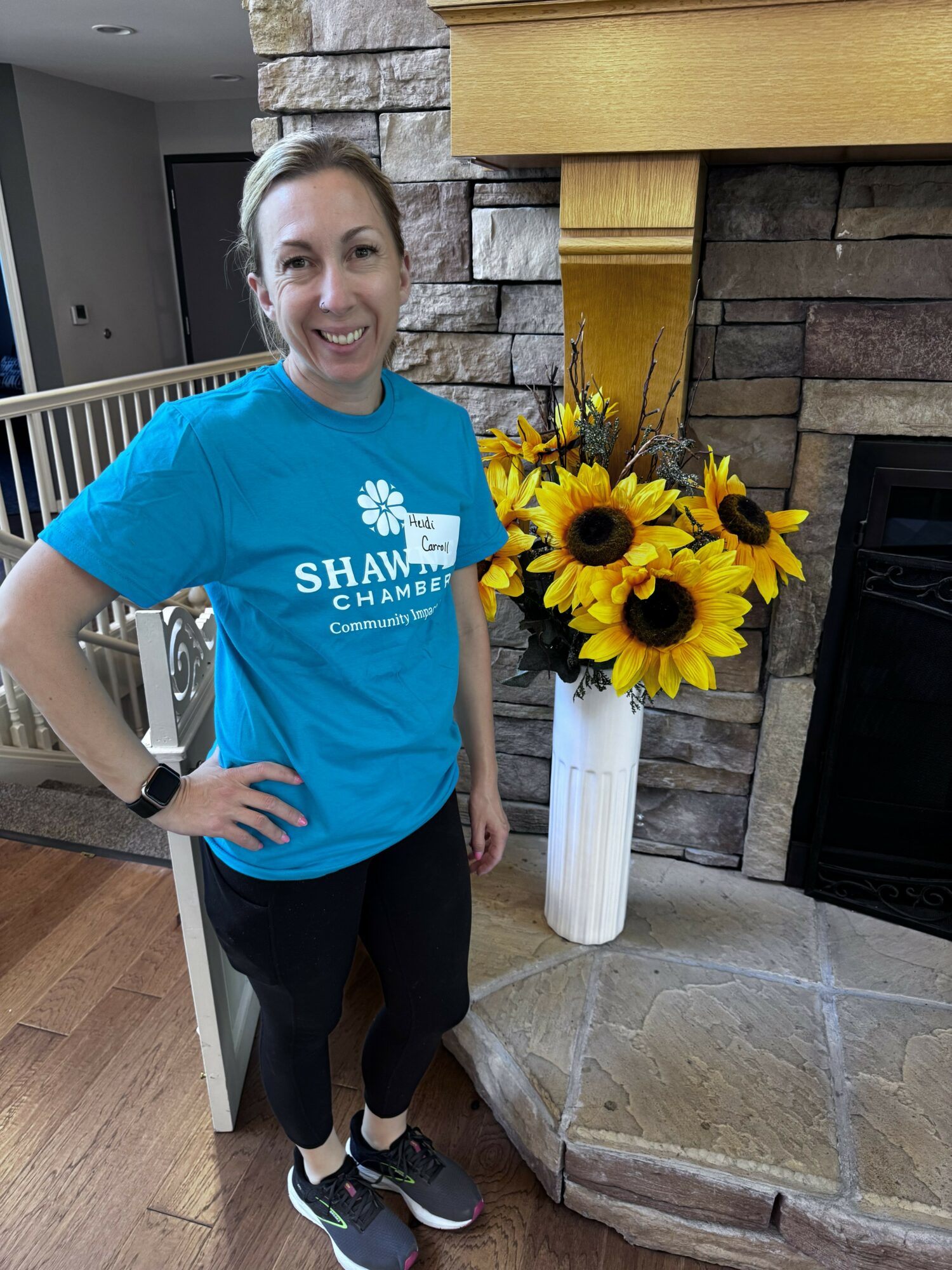 Woman in blue shirt standing next to a sunflower arrangement in a white vase near a fireplace.