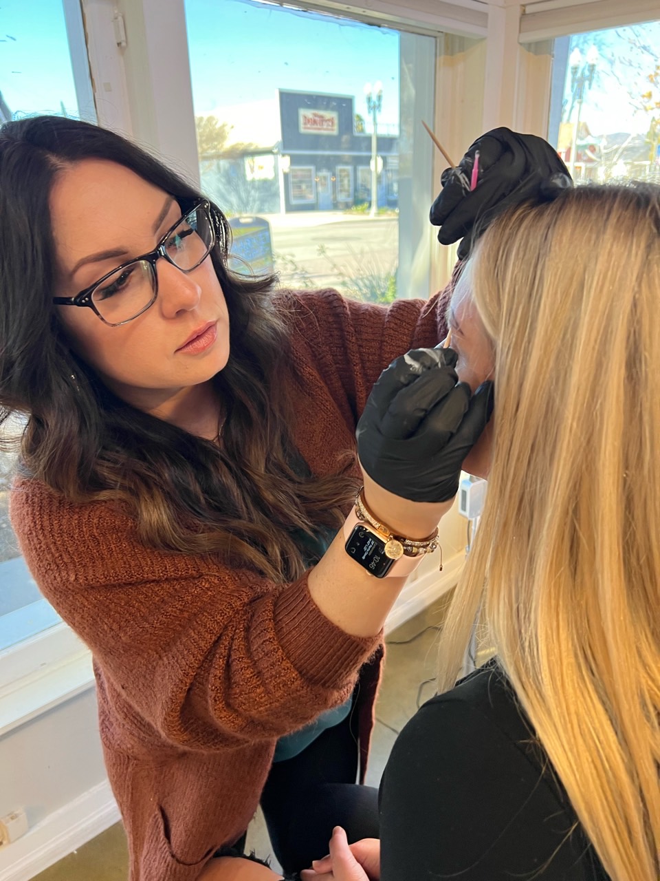 Woman with glasses and brown sweater applies makeup to blonde woman with black gloves inside a room with large windows.