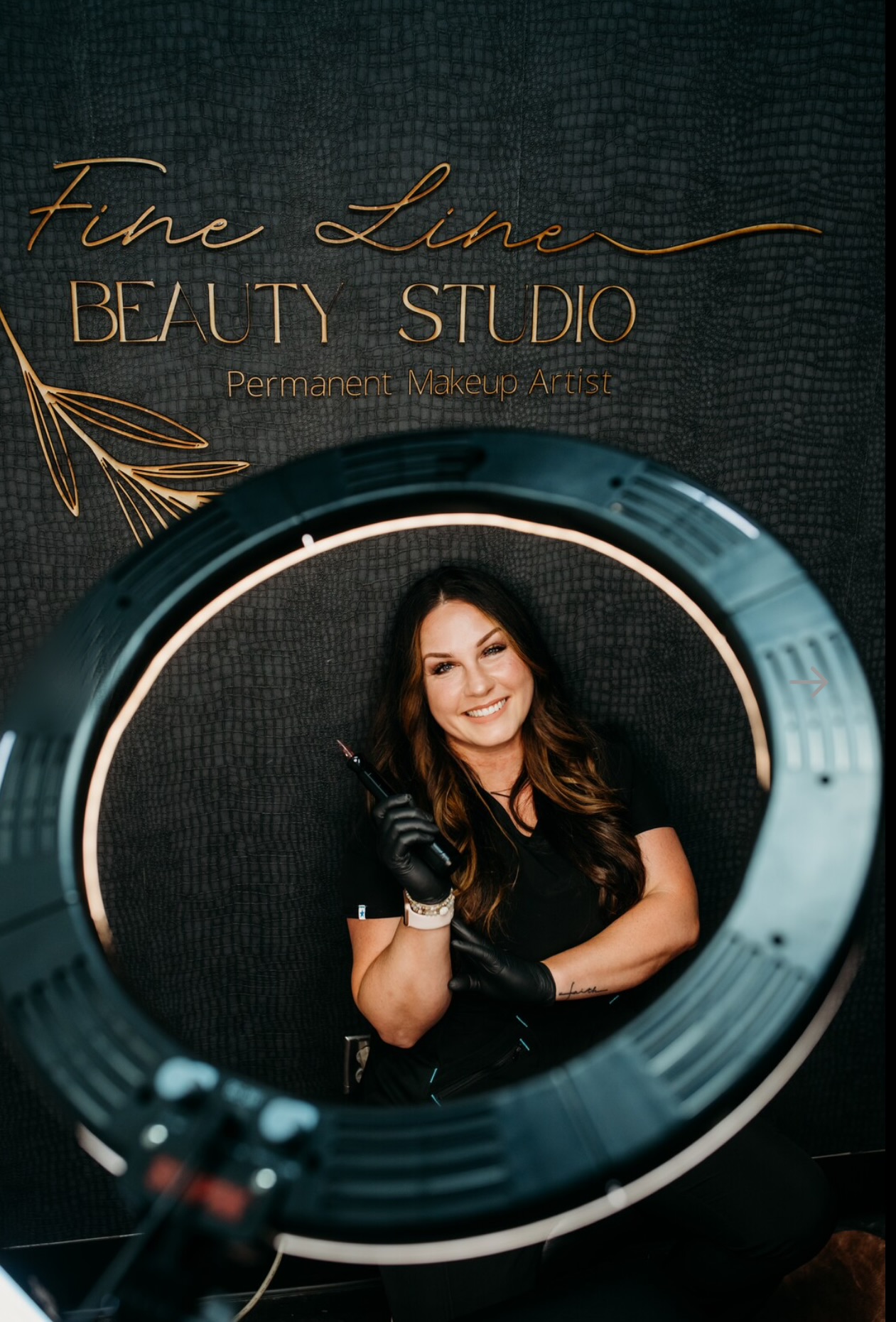 Smiling woman with long hair, wearing black gloves, sitting in a ring light at a beauty studio.