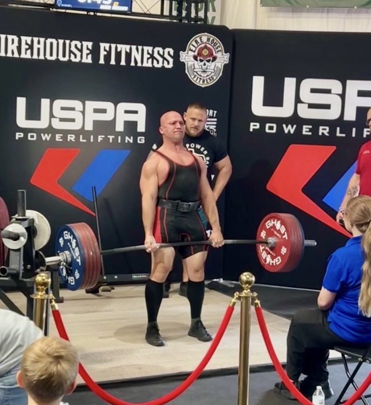 Powerlifter holding a barbell with weights, standing on stage with USPA and Firehouse Fitness banners, audience watching.