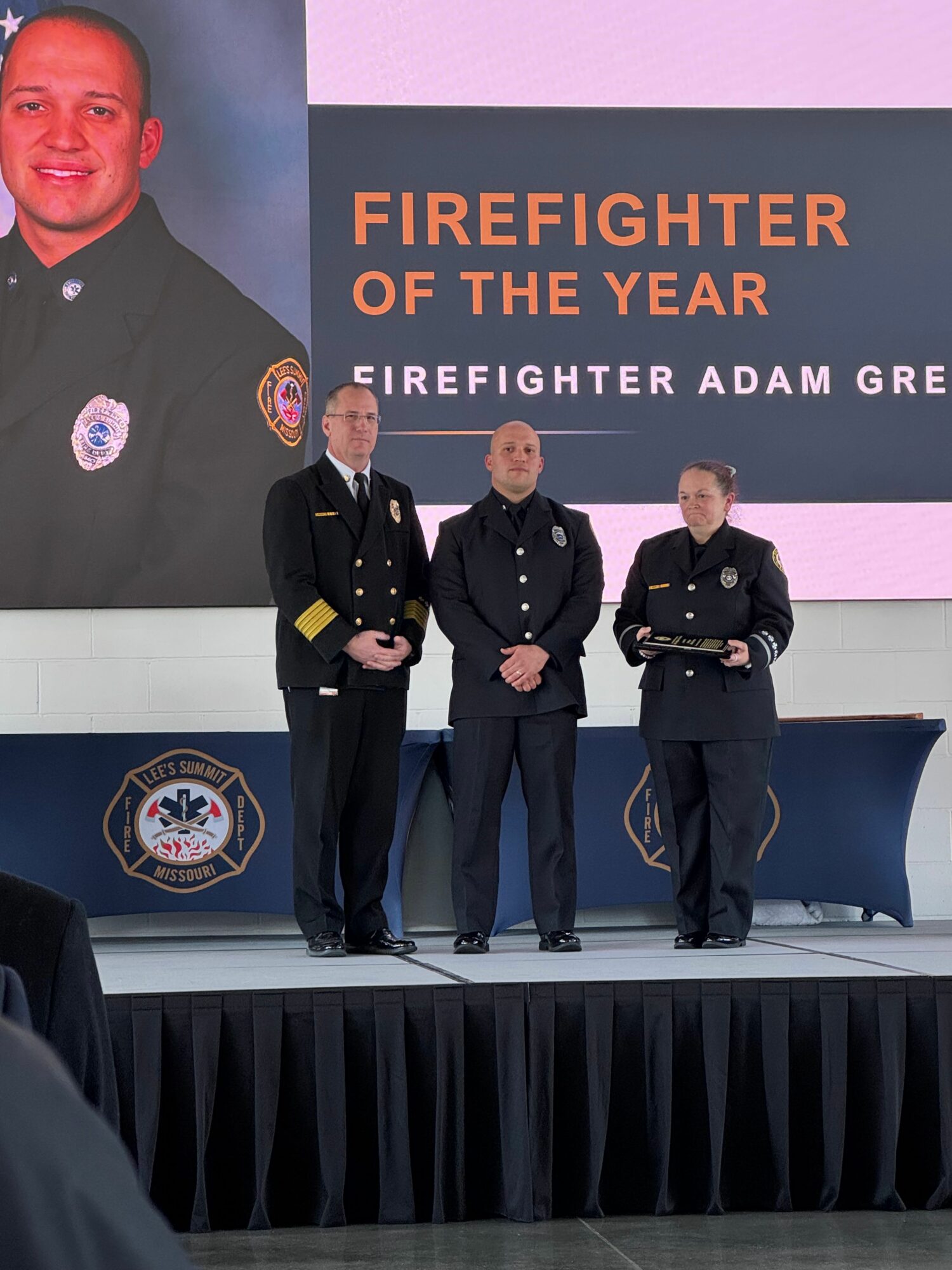 Three firefighters in uniform standing on stage during an award ceremony, with a large screen behind them displaying a firefighter's photo and text.