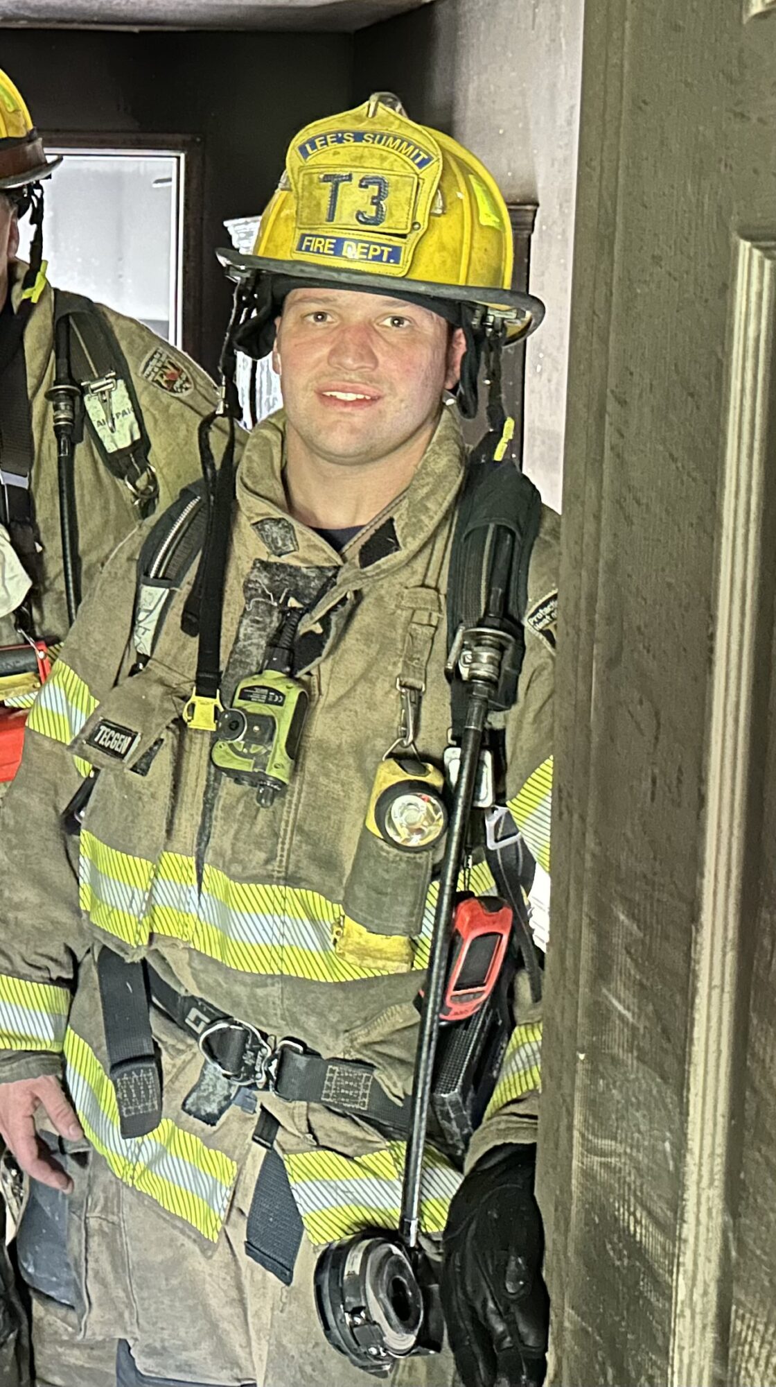 Firefighter in uniform and helmet standing indoors, smiling, with equipment and a wall behind.