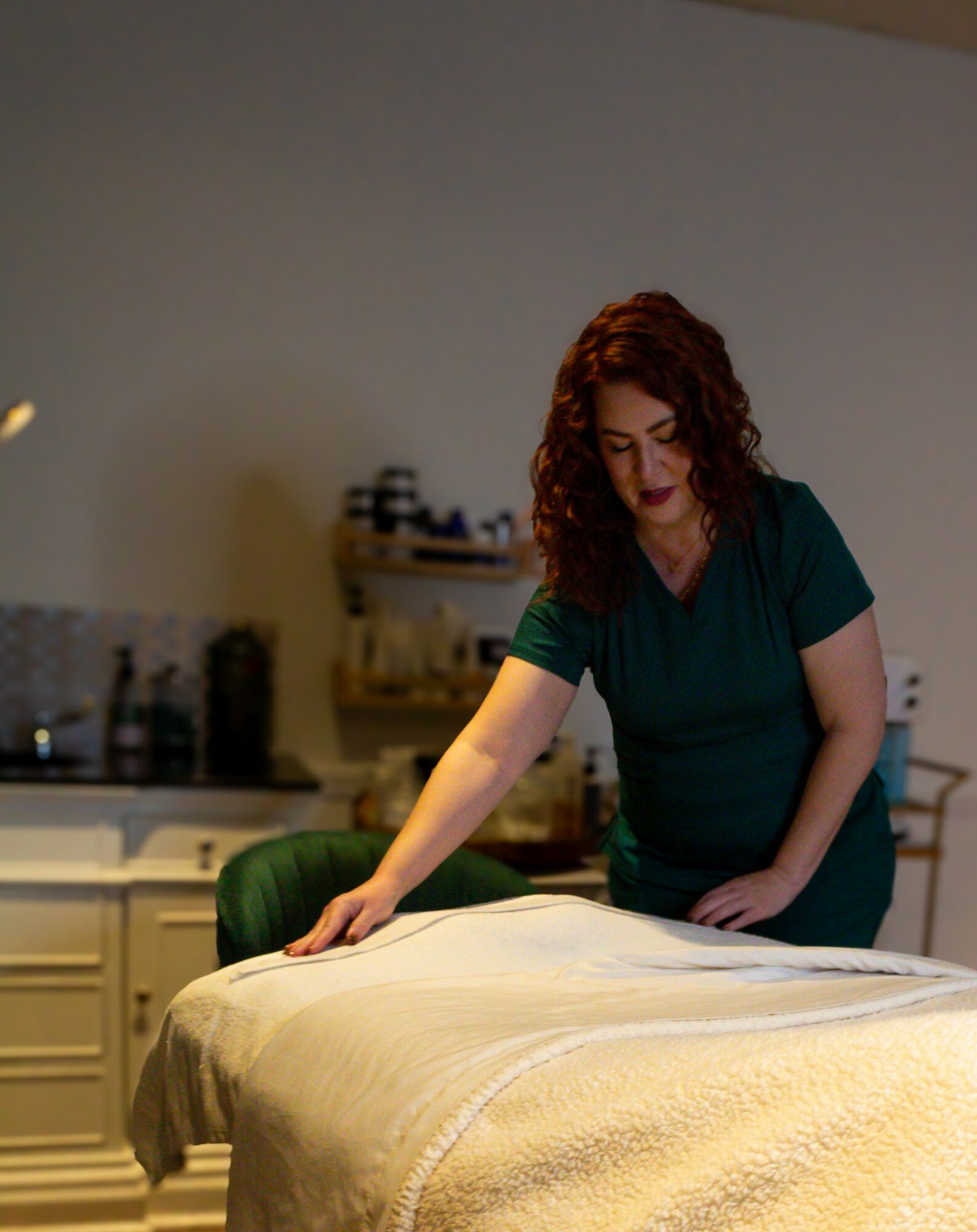 Woman with curly hair leaning over a massage table in a room with shelves and equipment in the background.