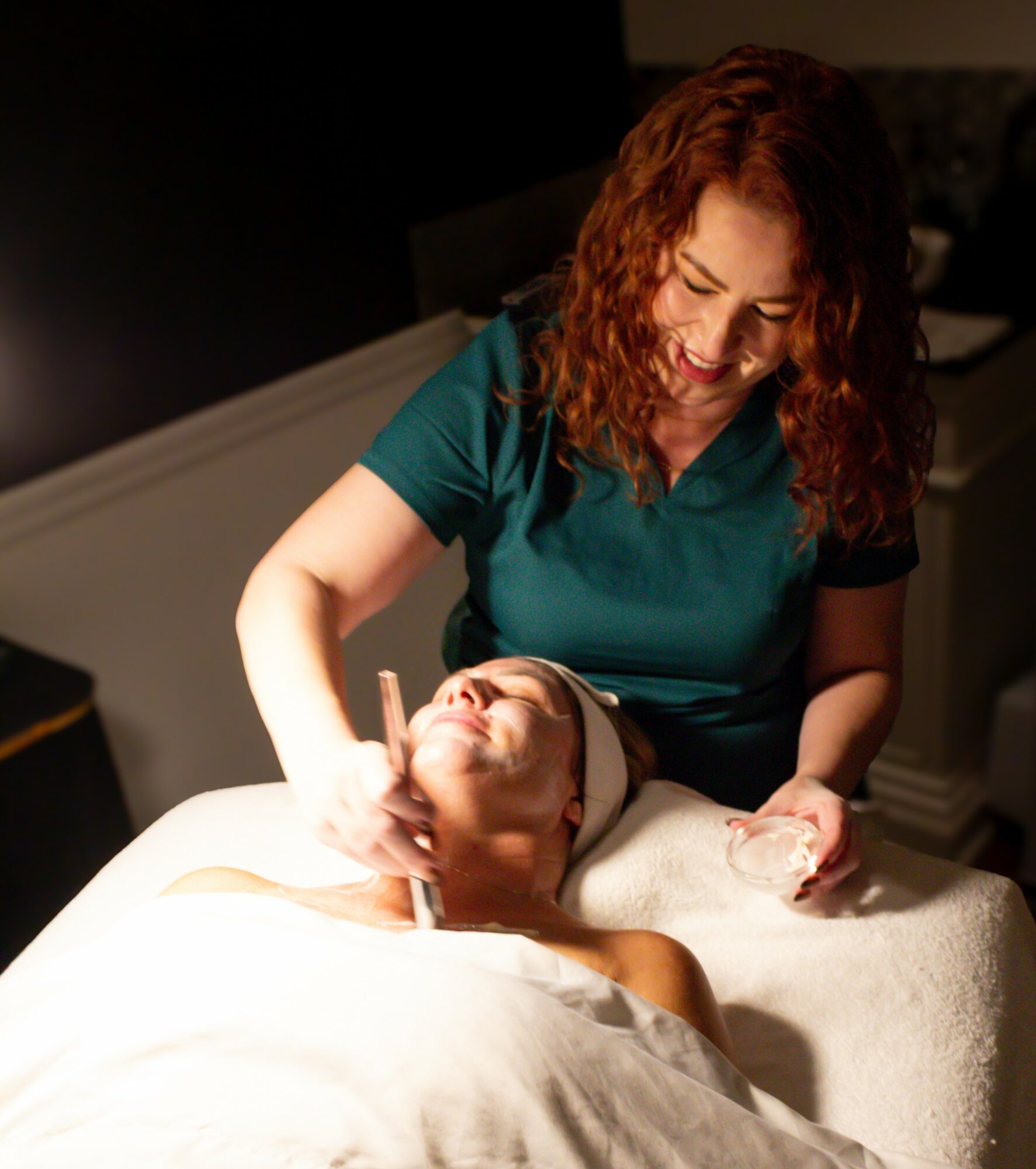 Woman with curly hair performs facial treatment on a person lying down, in a dimly lit room.