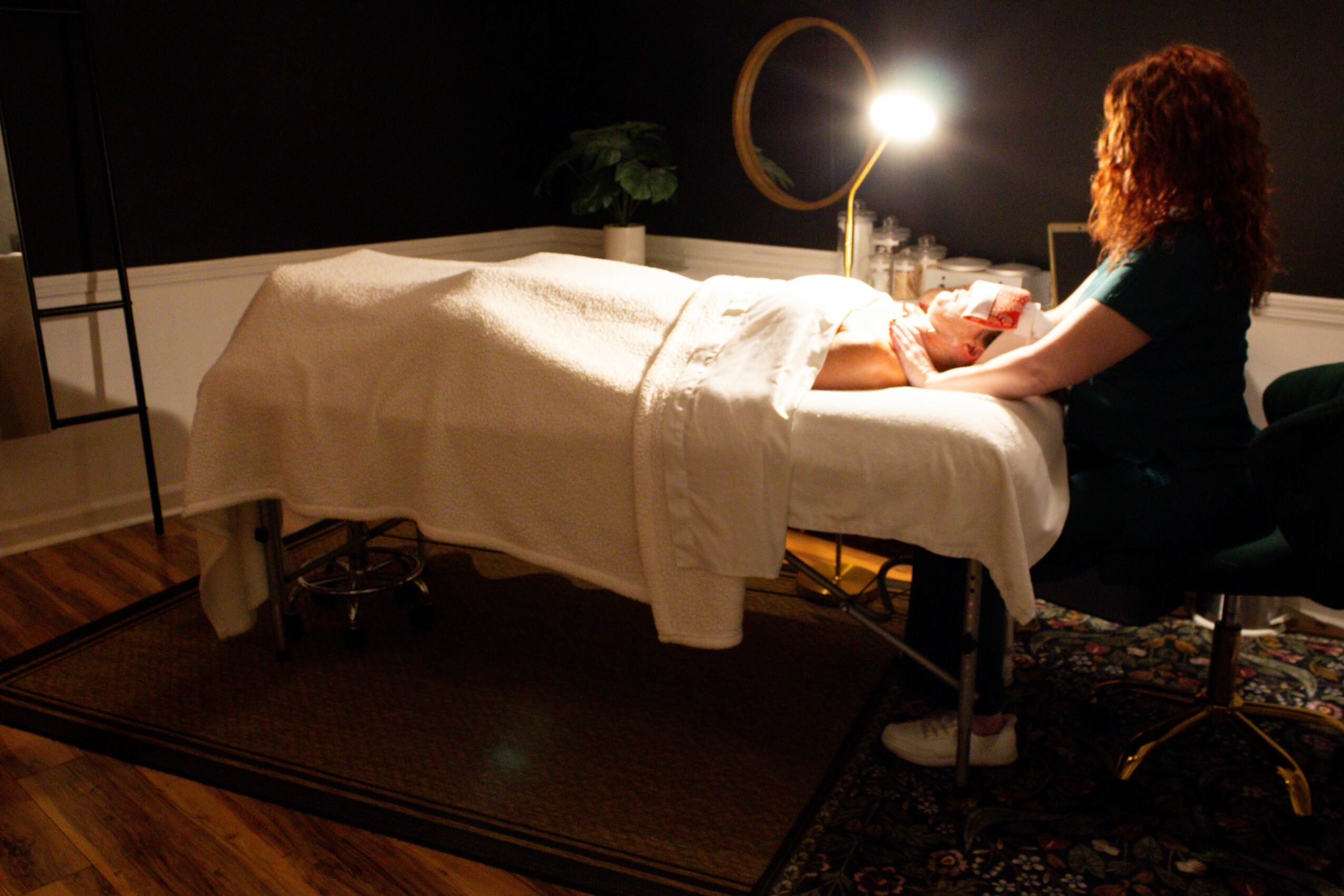 A woman with curly hair sits beside a bed in a dimly lit room, holding a small object, with a lamp nearby.